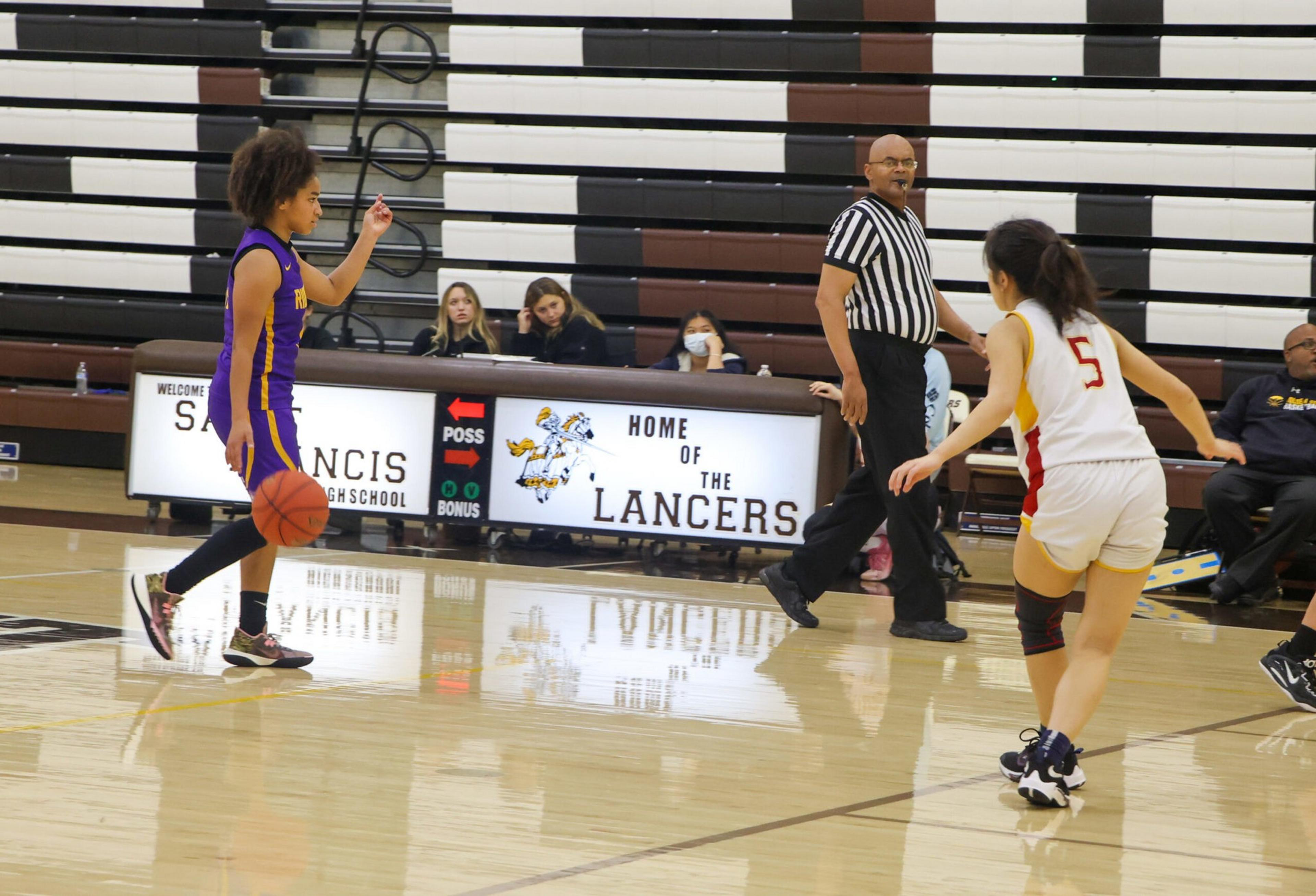 A basketball player in purple dribbles the ball while being guarded by a player in white with number 5, with a referee walking nearby and spectators seated.