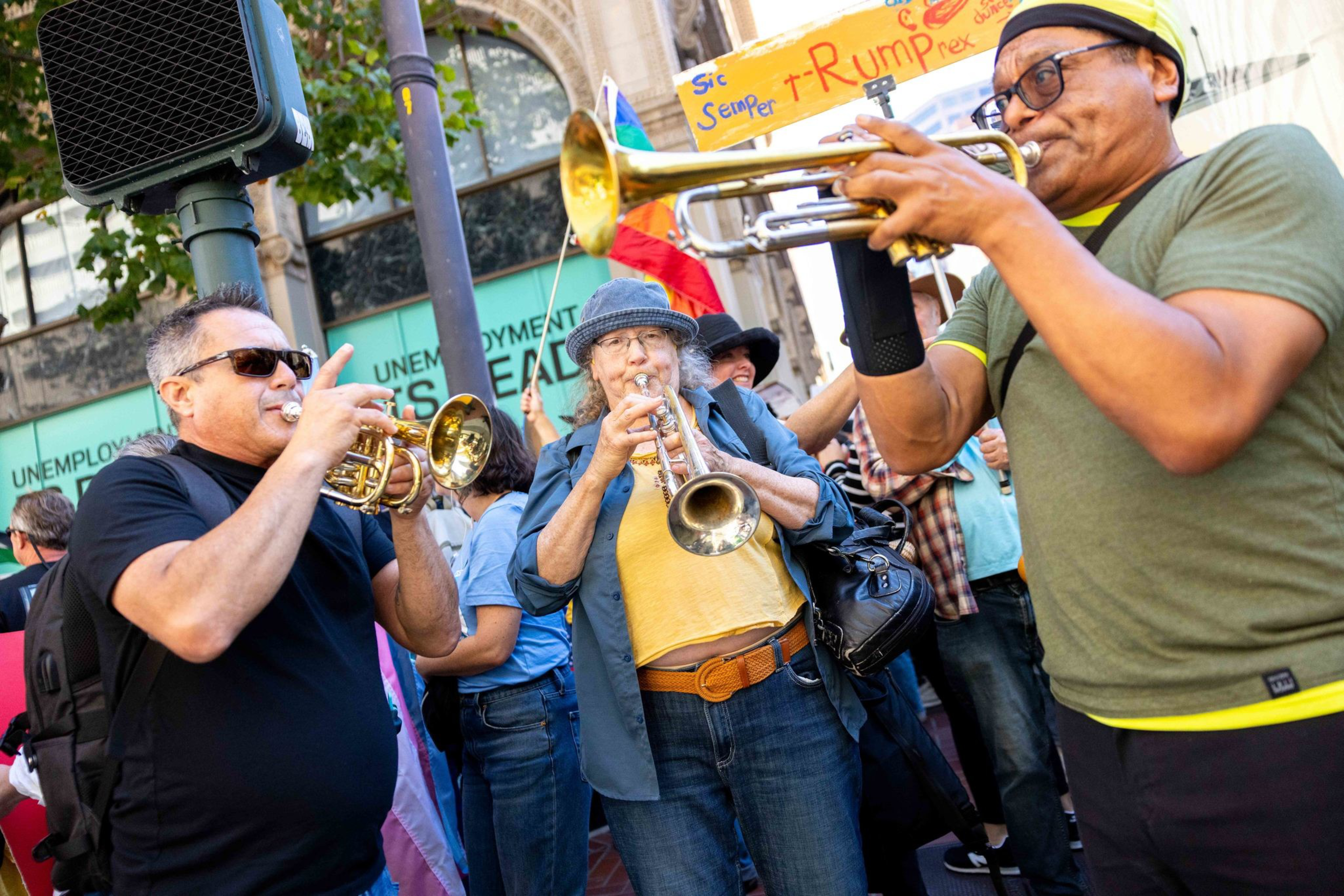 Three people playing trumpets outdoors in a crowd, with signs and a building in the background.