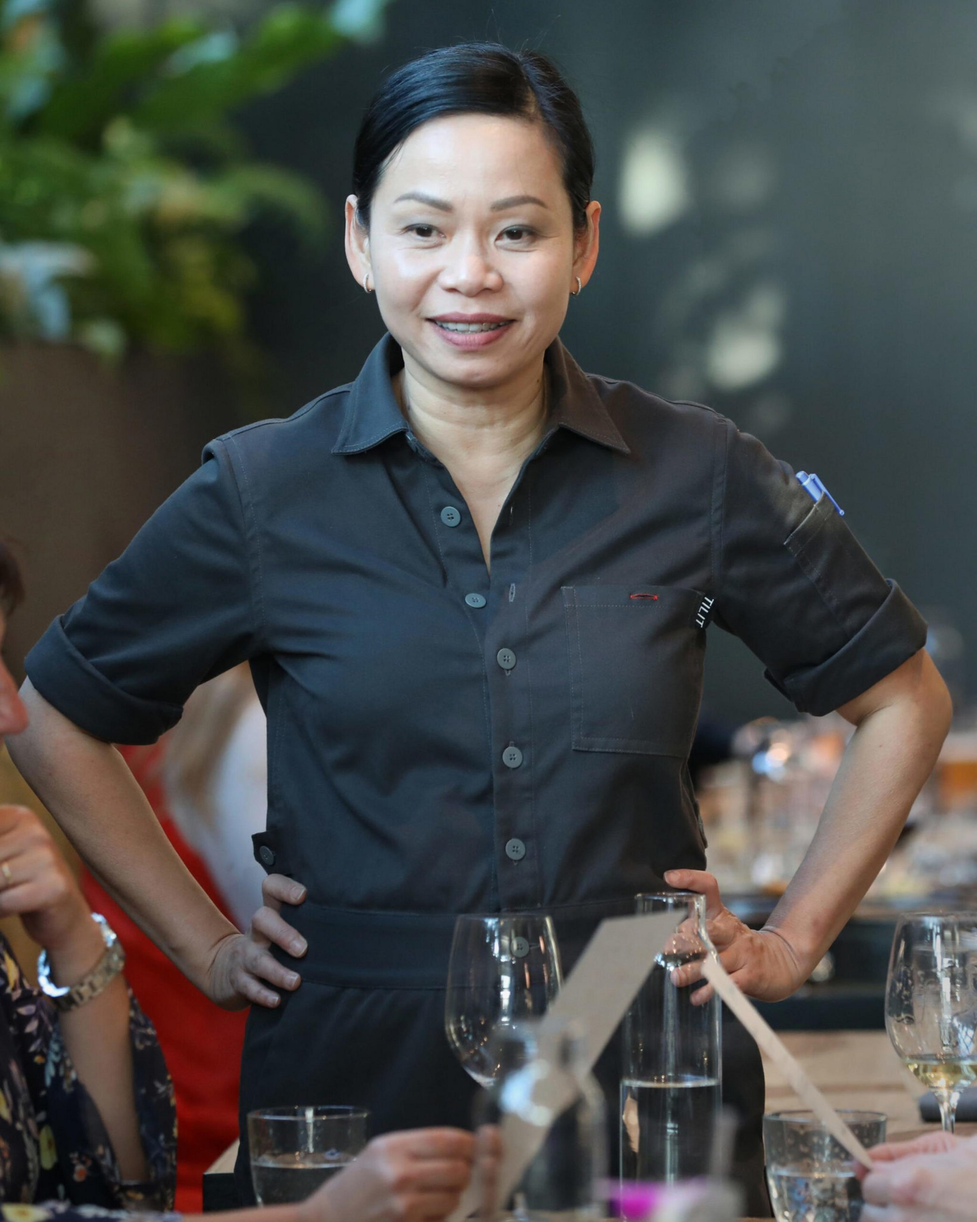 A woman in a dark, short-sleeved shirt stands with her hands on her hips, smiling at a table with glasses and menus. There are blurred plants in the background.