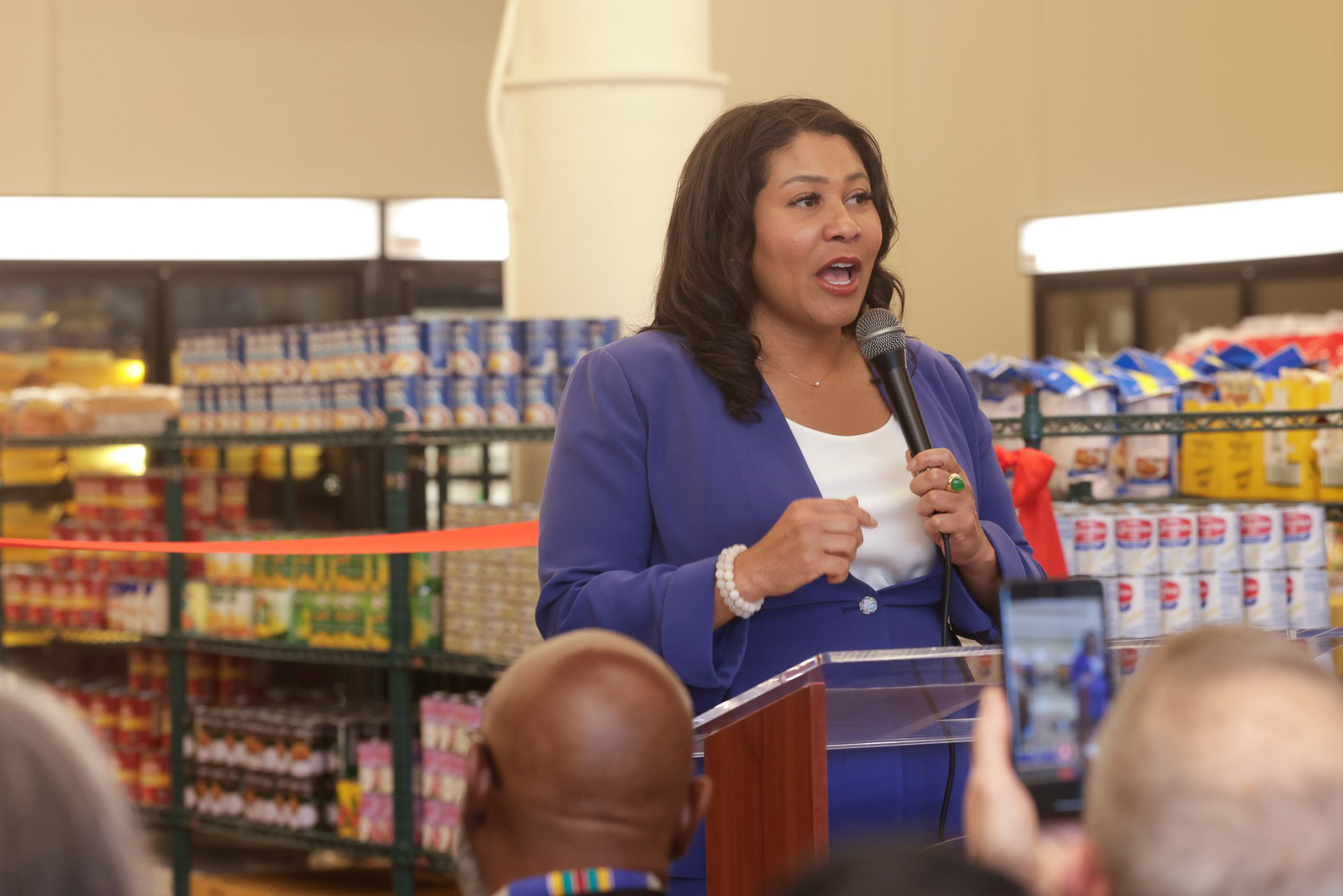 A woman in a blue suit speaks into a microphone in front of a grocery store shelf while people in the foreground watch and record her with a smartphone.