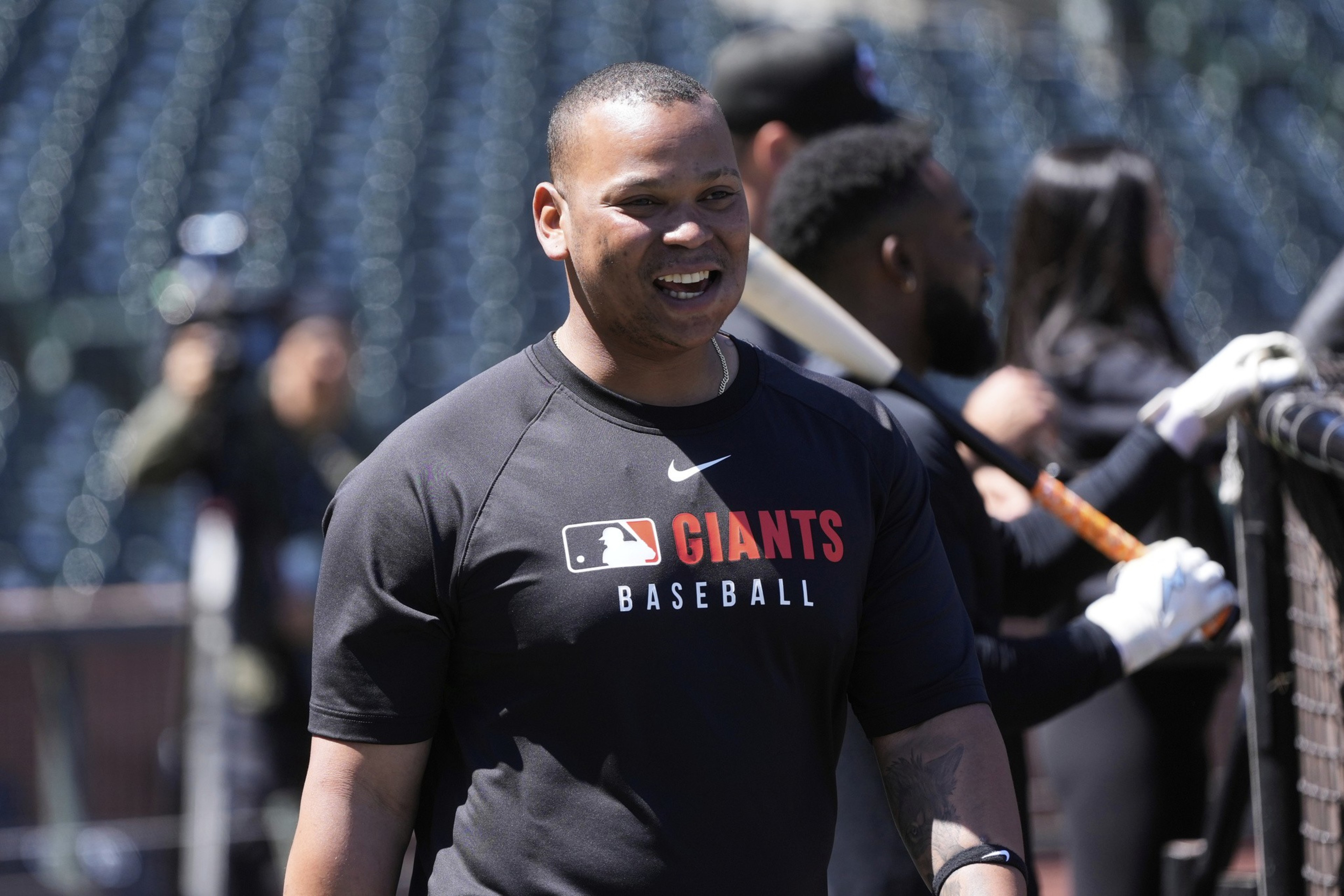 A smiling person in a &quot;Giants Baseball&quot; shirt stands in a stadium, with blurred figures in the background holding baseball bats and equipment.