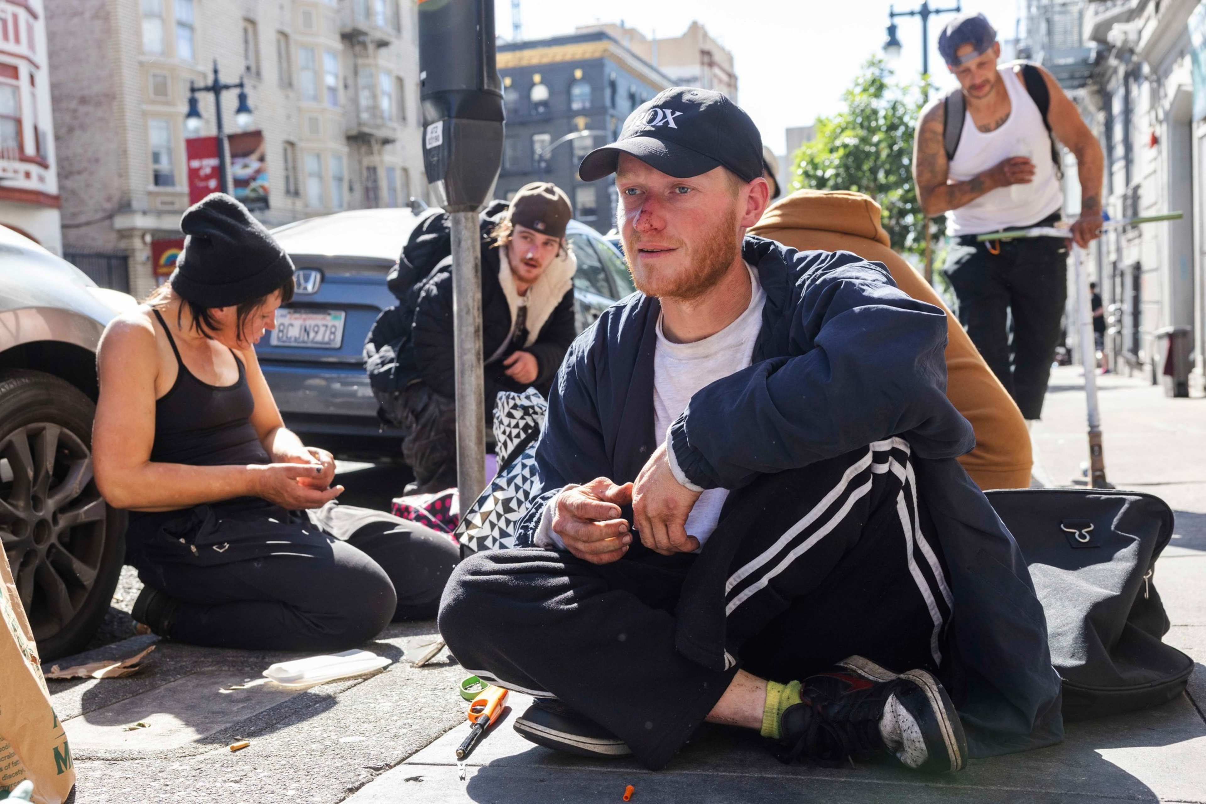 Four people sit or crouch on a city sidewalk, with one person in a ...
