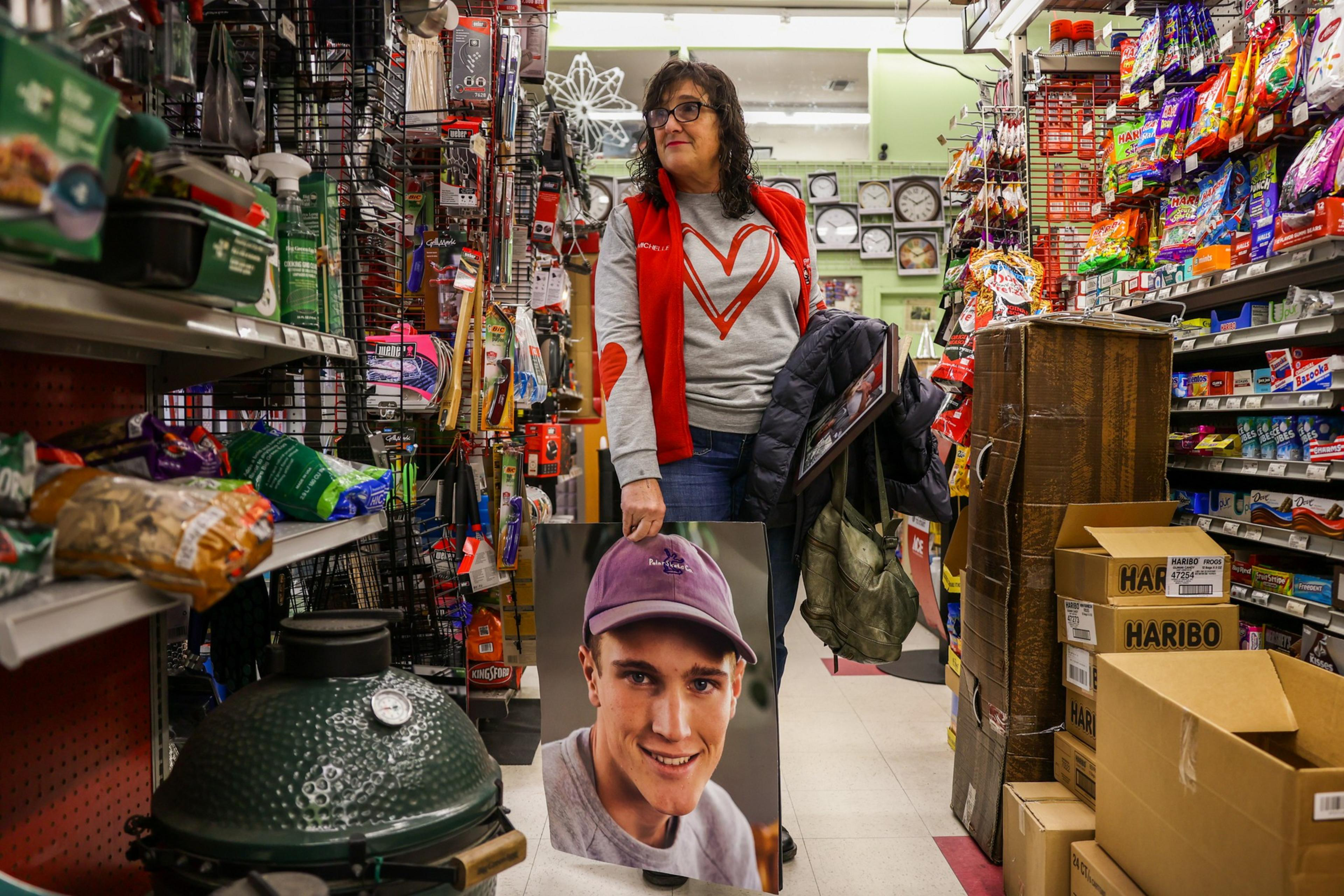 A woman stands in a store aisle holding a large photo of a smiling young man wearing a purple cap, surrounded by shelves filled with various products.