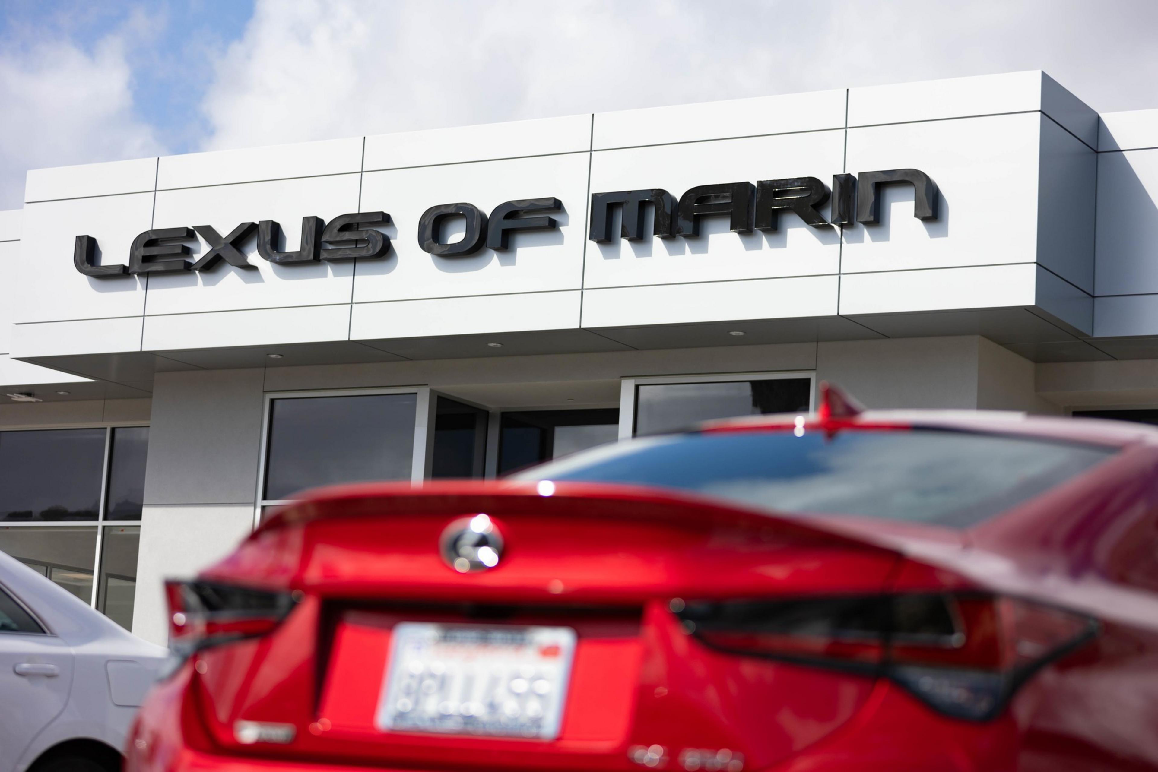A Lexus dealership named "Lexus of Marin" is shown. In the foreground, there's a red Lexus car facing away. The sky is partly cloudy above the building.