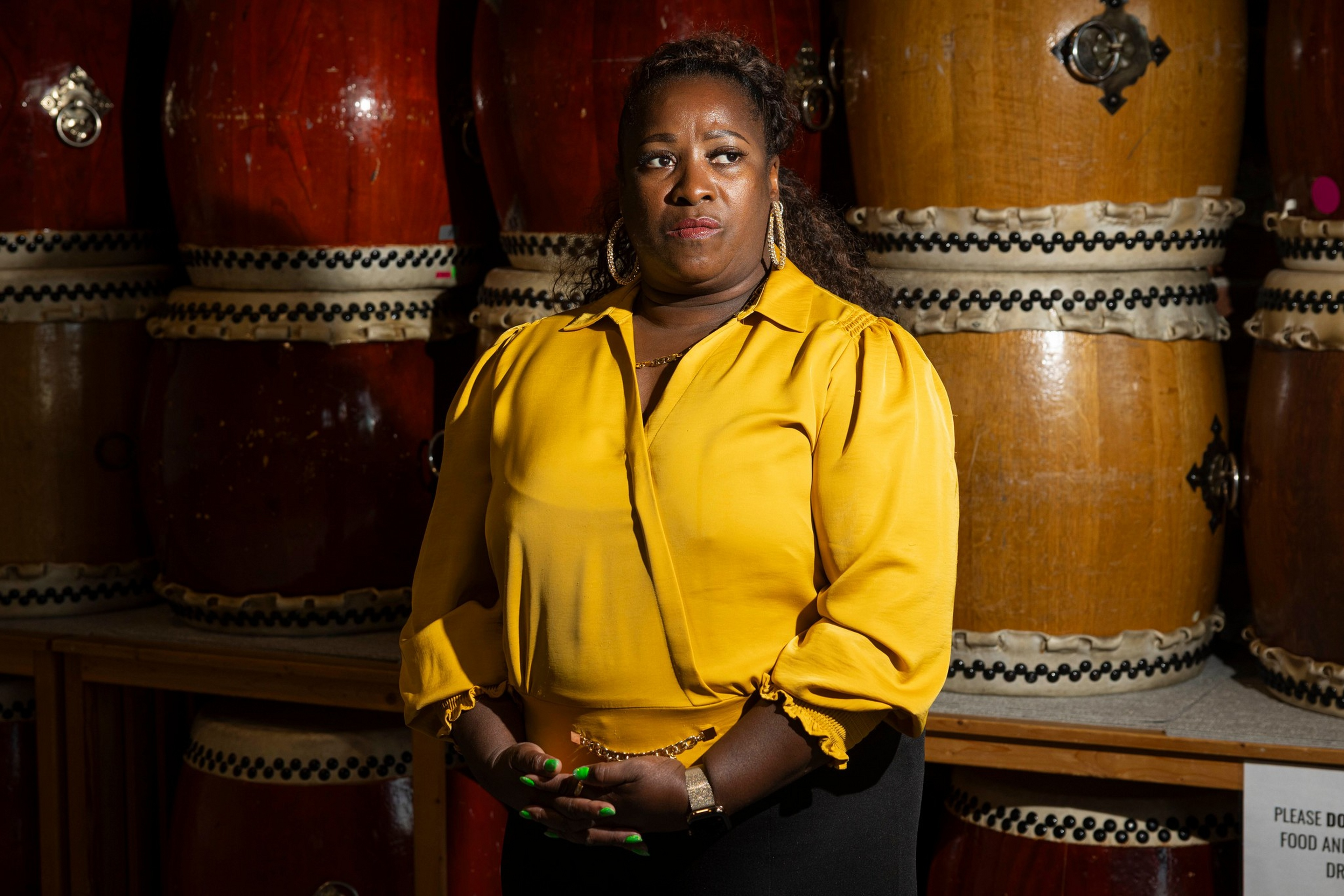A woman in a yellow blouse stands with clasped hands in front of large traditional drums, looking to the side with a serious expression.