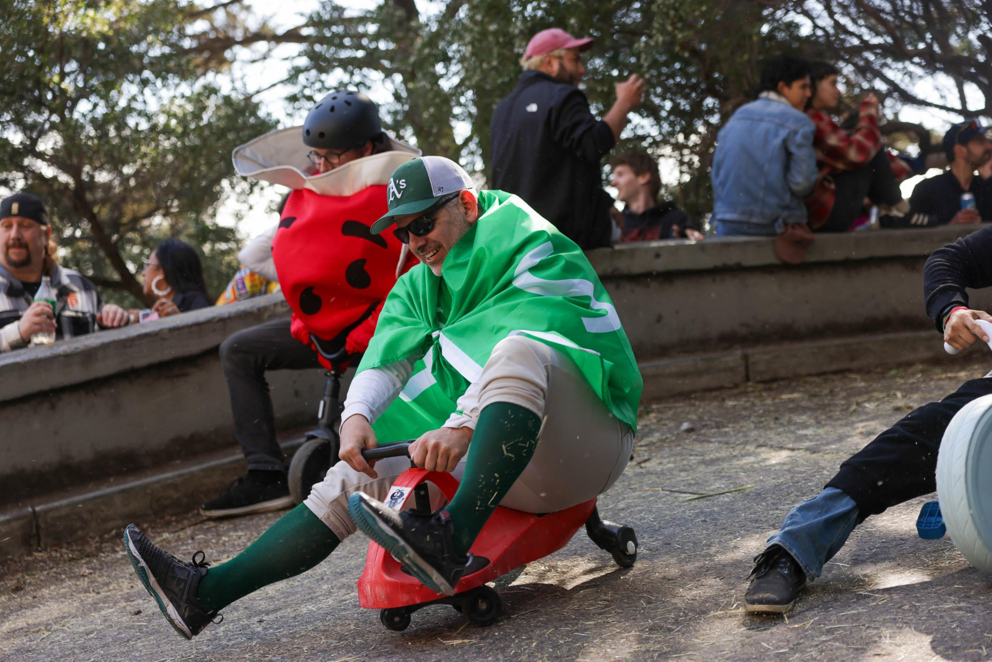 People in costumes ride small plastic scooters downhill, spectators watch in the background.