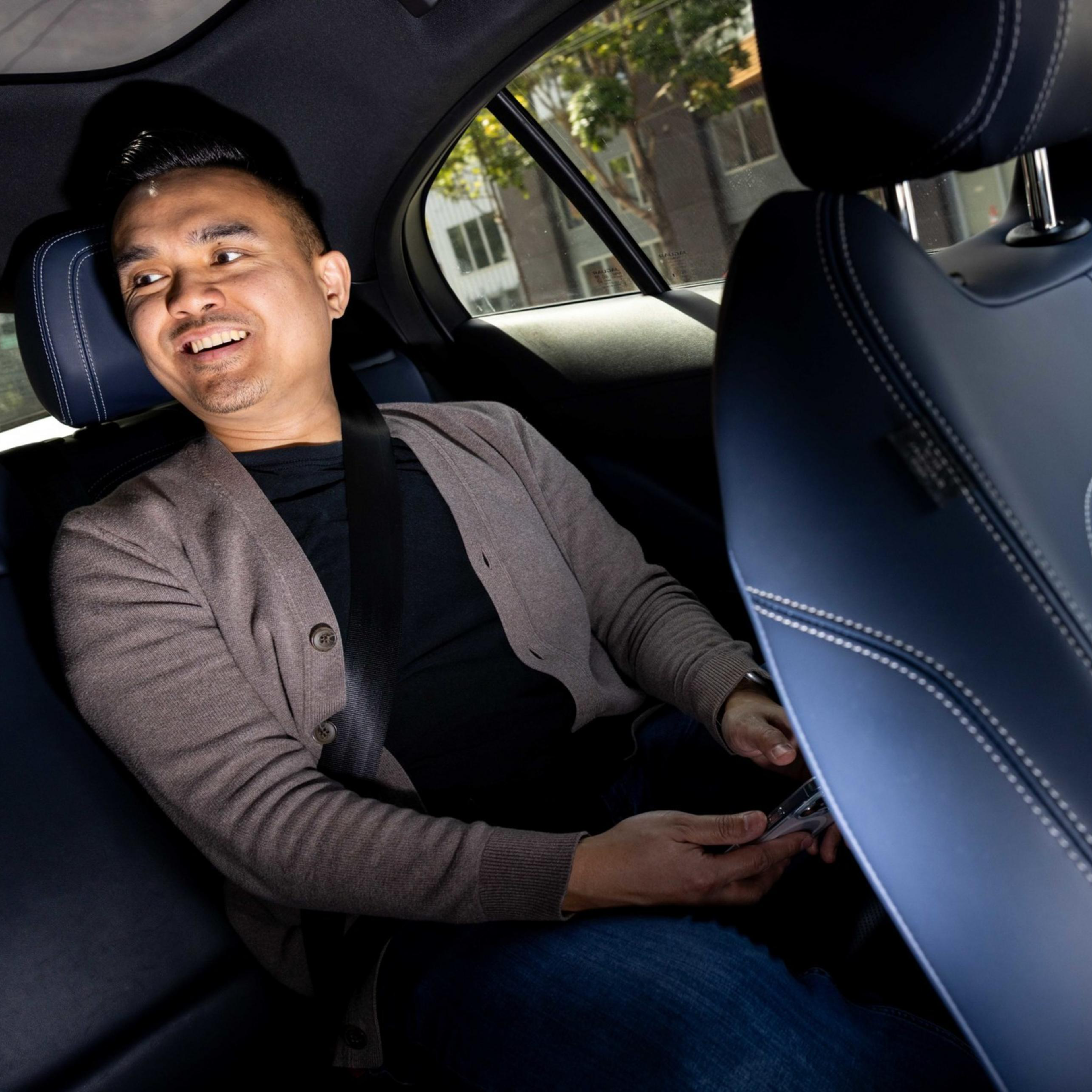 A person wearing a gray cardigan is smiling while seated in the backseat of a car, holding a phone. The car interior is dark with blue accents.