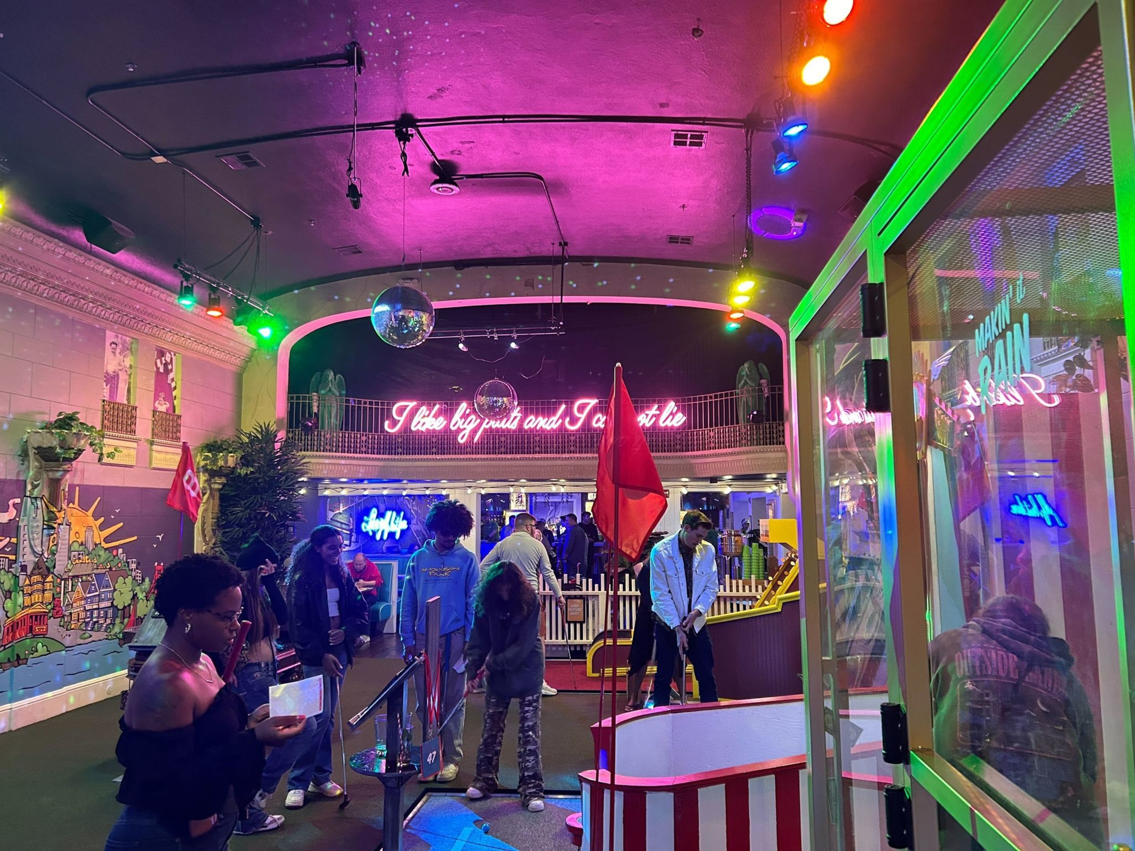 A colorful, lively indoor mini-golf venue with neon signs, people playing, and vibrant murals. There are disco balls and multi-colored lights, creating a fun, upbeat atmosphere.