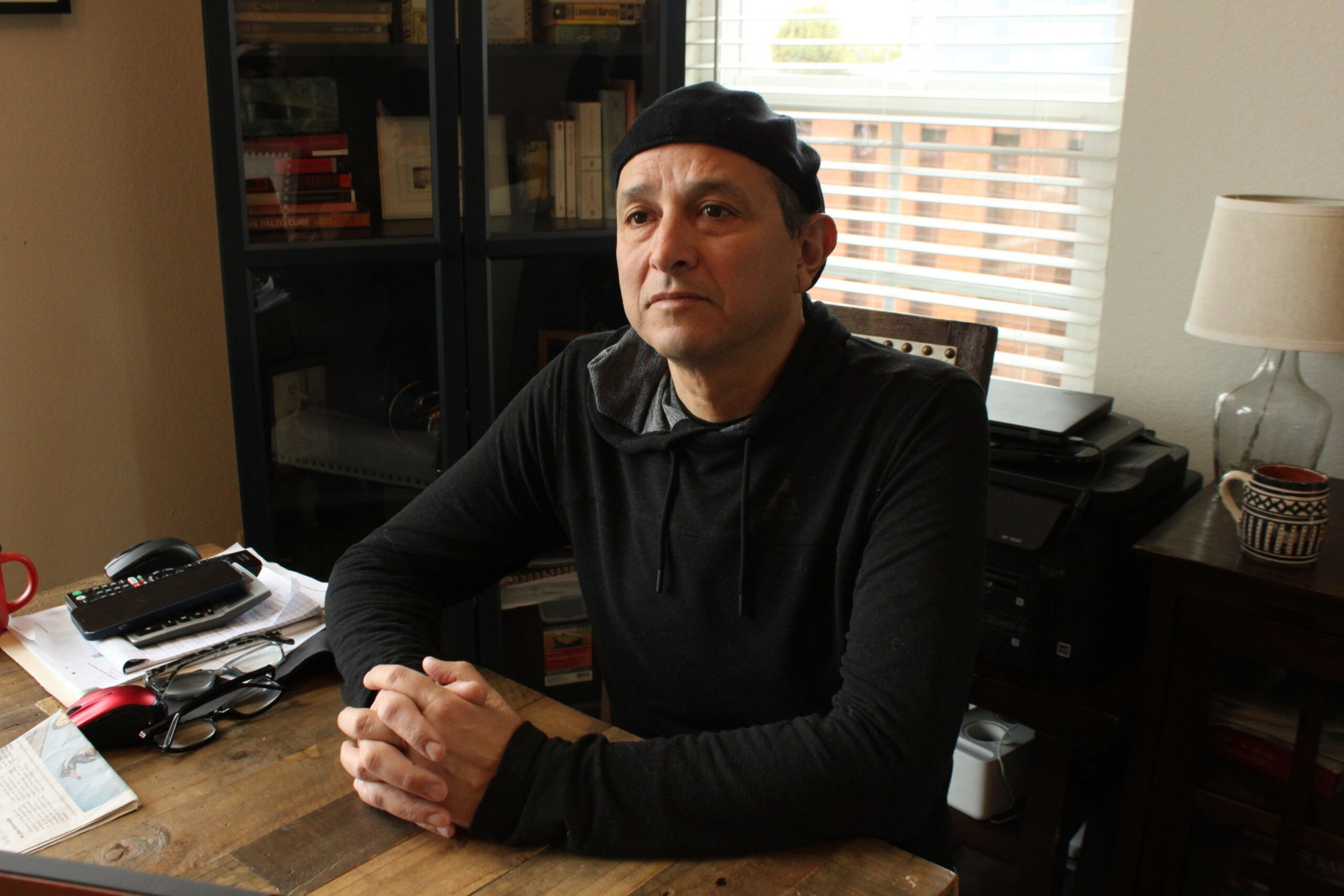 A man wearing a black hoodie and beret sits at a wooden table with clasped hands, looking thoughtful, with a bookshelf and window behind him.