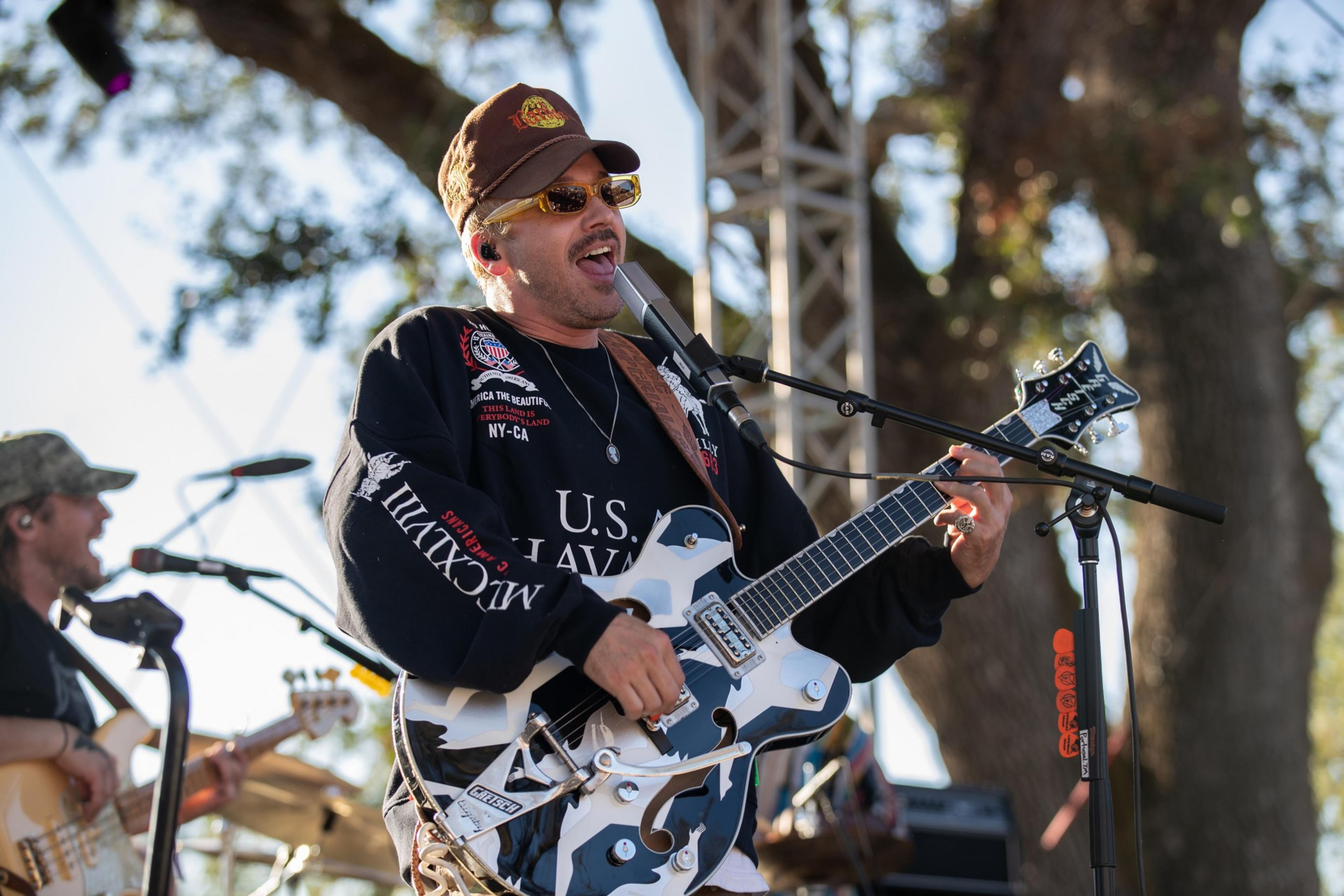 A man wearing sunglasses, a cap, and a black "U.S. Navy" sweatshirt sings and plays a camouflage-patterned electric guitar on an outdoor stage.