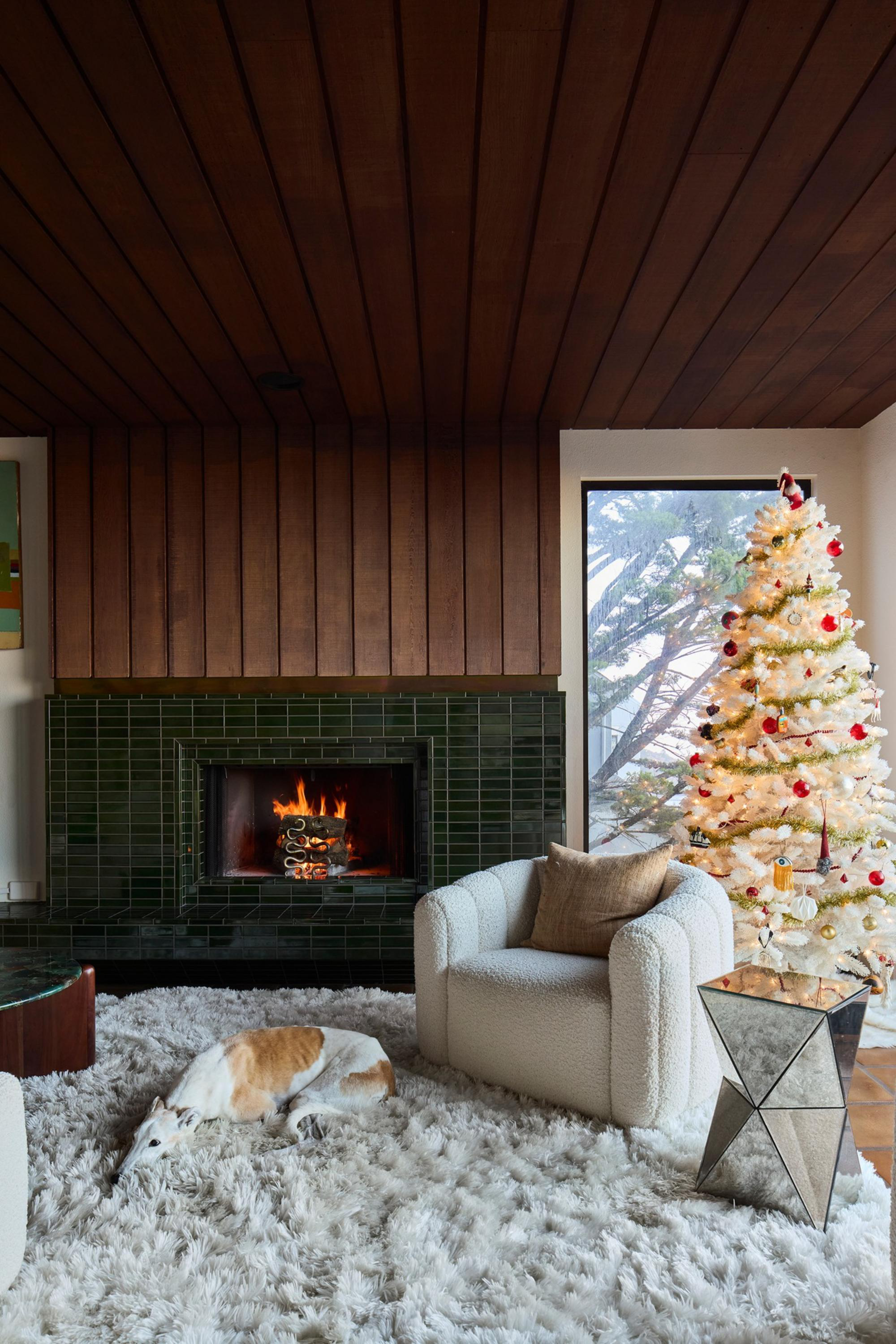 A cozy living room features a lit fireplace, a fluffy white rug with a resting dog, a cushioned armchair, a mirrored side table, and a decorated white Christmas tree.