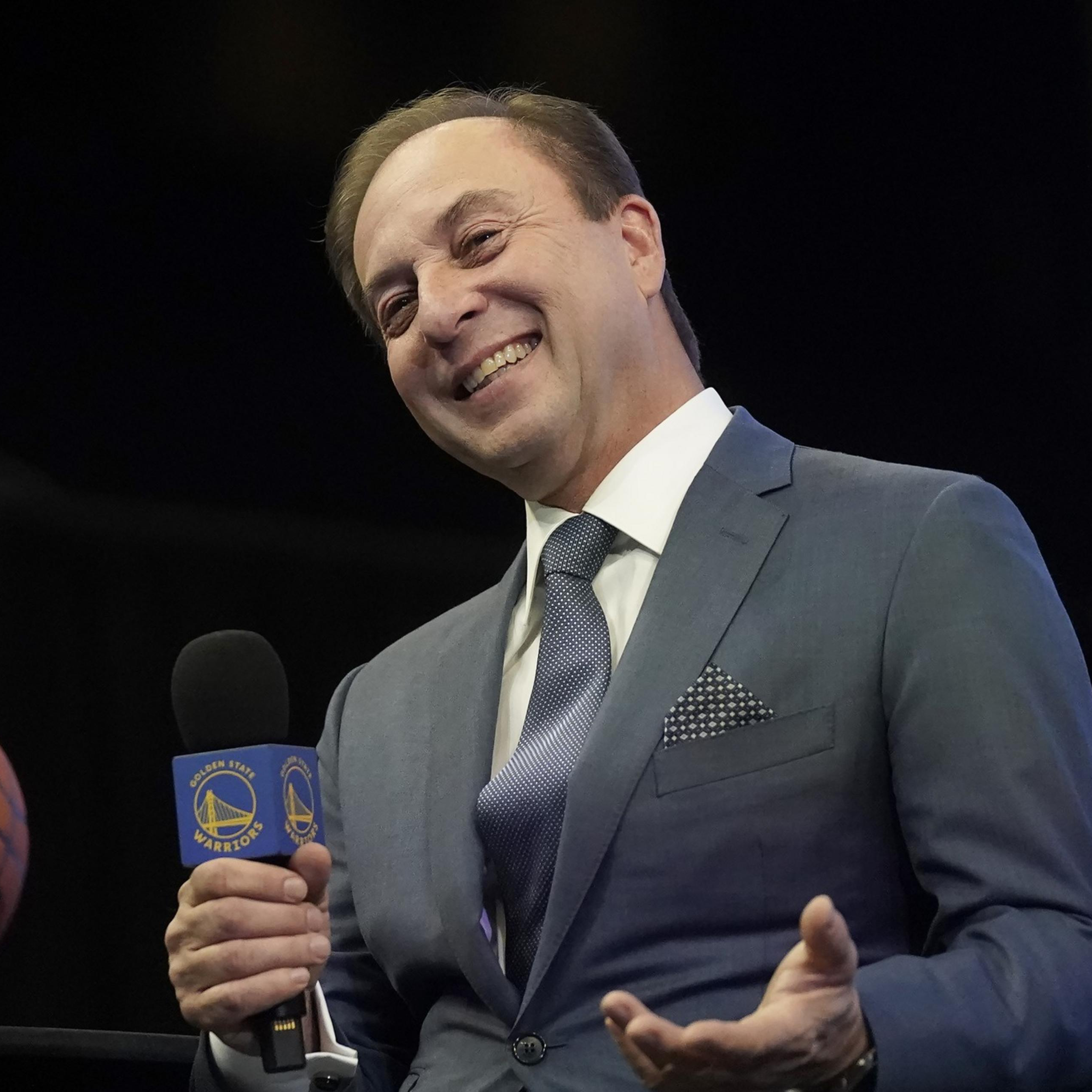 A man in a suit smiles while holding a microphone with a Golden State Warriors logo, with a basketball visible in the background.