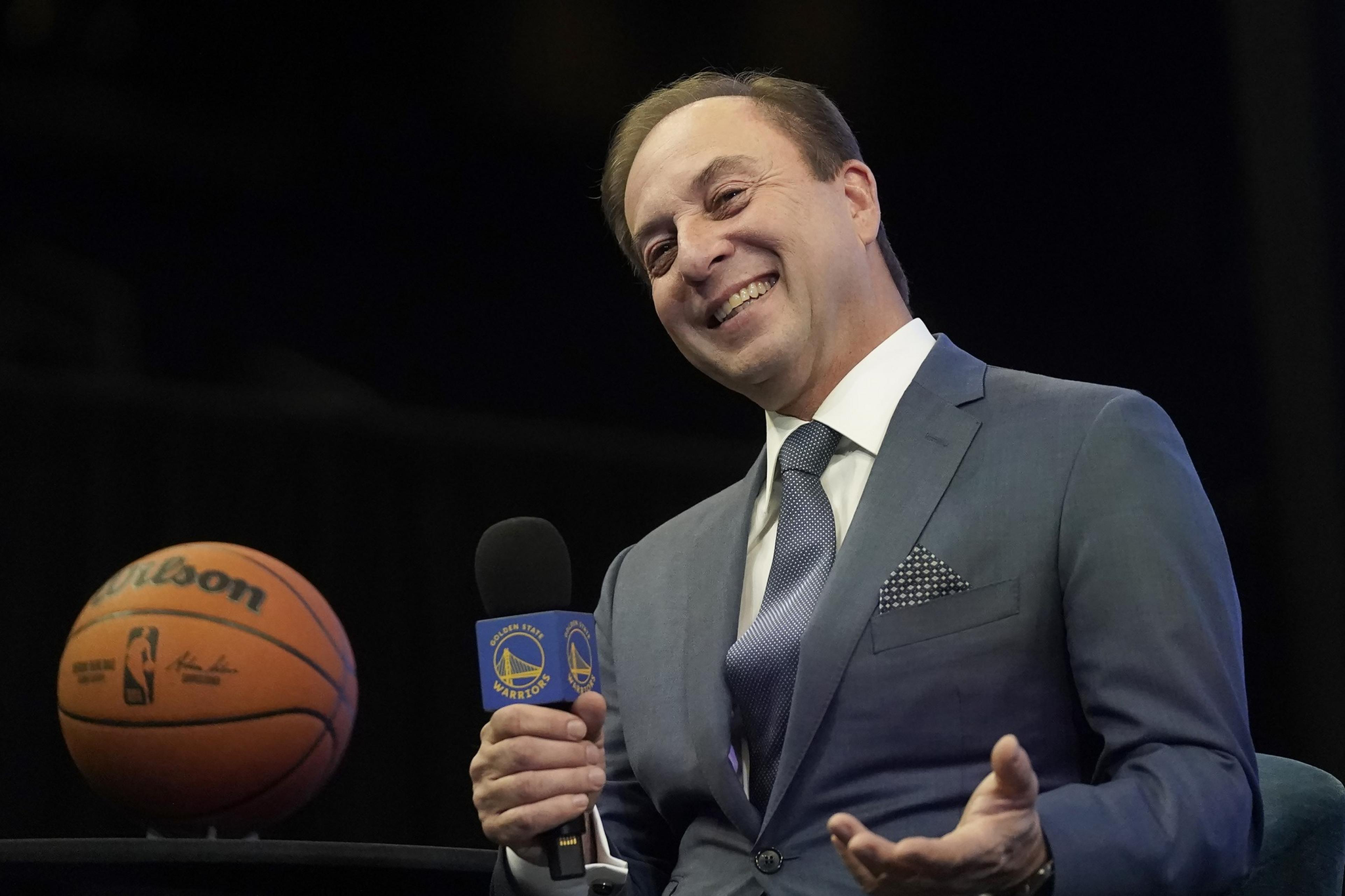 A man in a suit smiles while holding a microphone with a Golden State Warriors logo, with a basketball visible in the background.