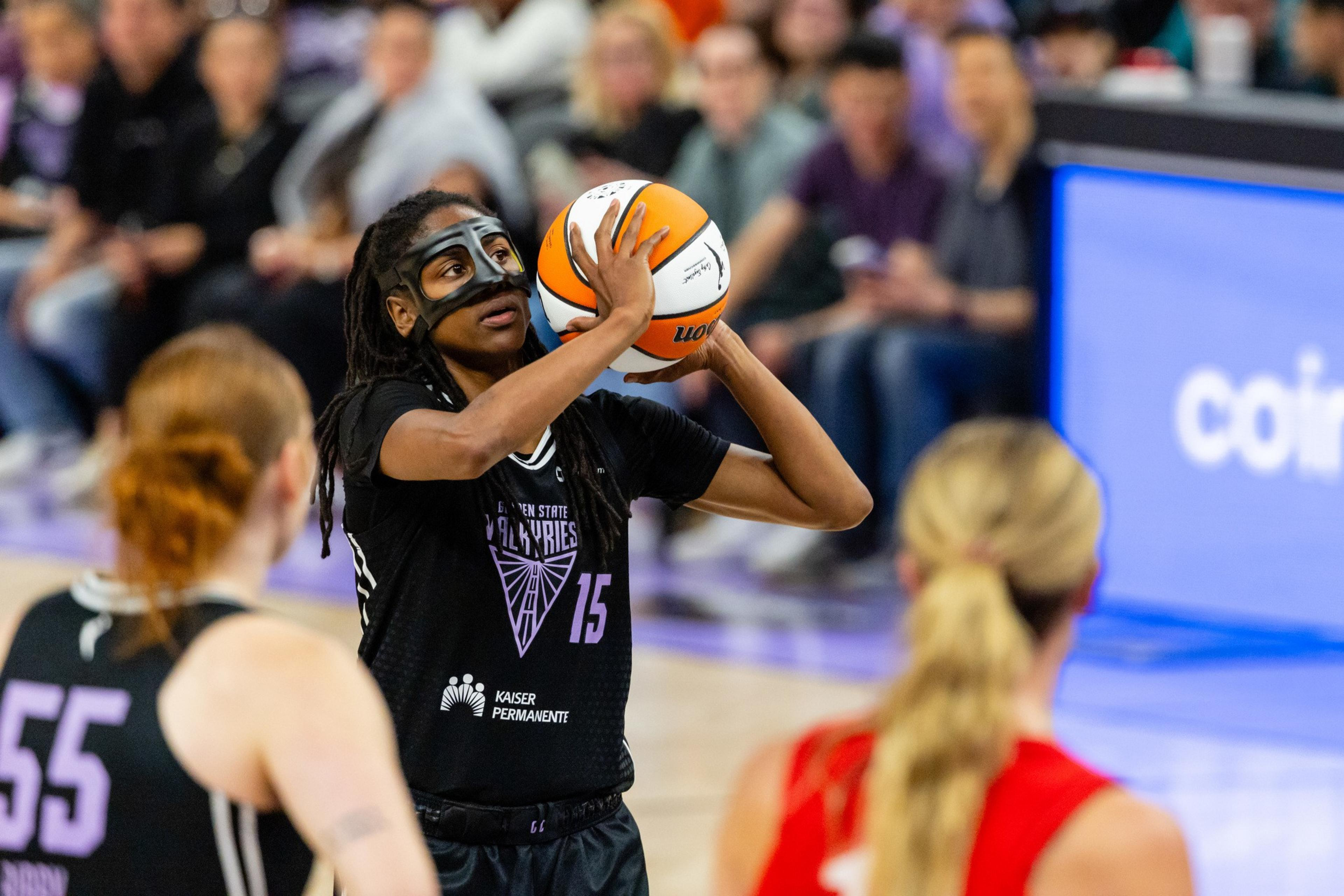 A basketball player wearing a protective mask prepares to shoot the ball. She's focused, with braided hair and black attire, while spectators watch in the background.