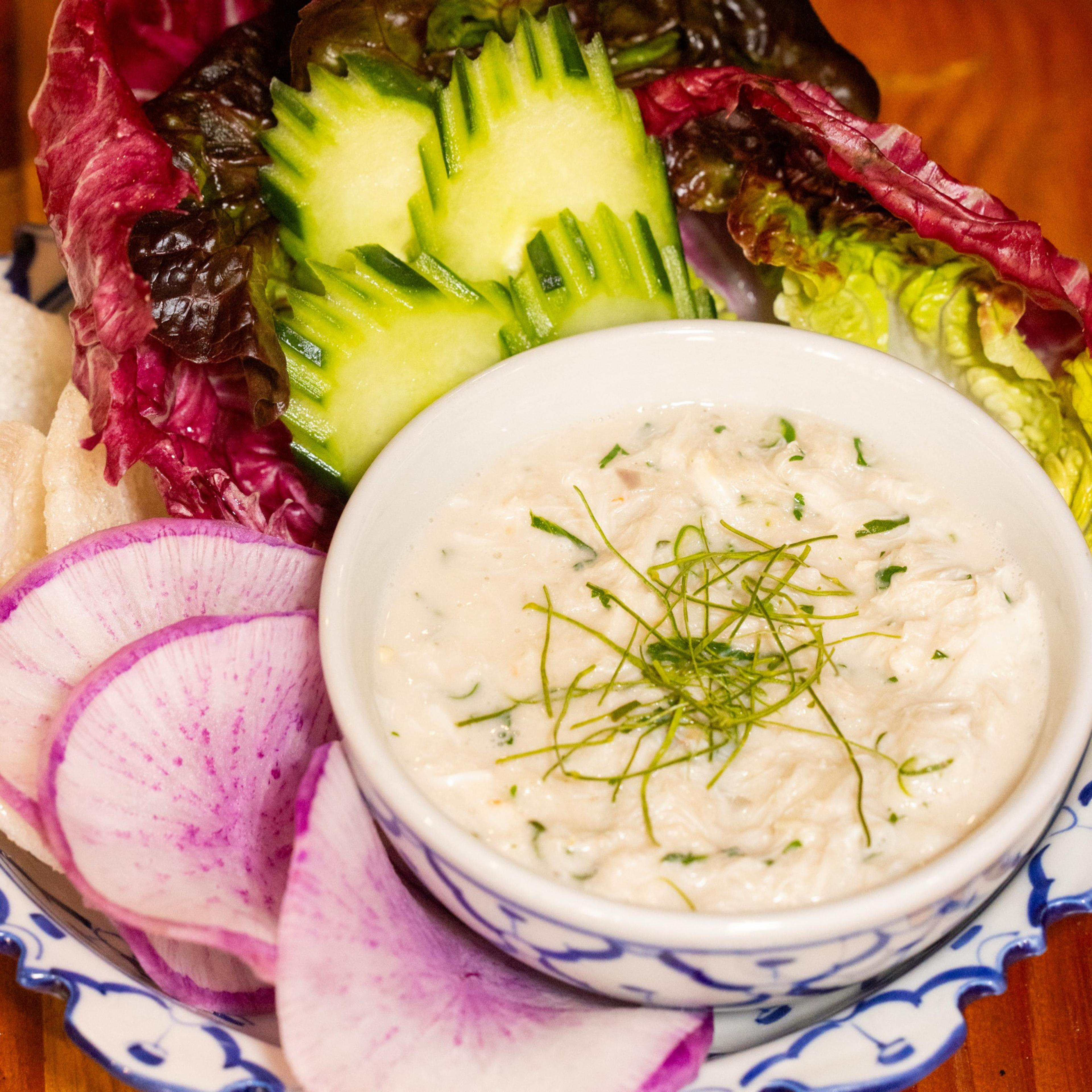 A bowl of creamy dip topped with herbs is surrounded by sliced radishes, cucumbers, and leafy greens on a decorative blue-and-white plate.