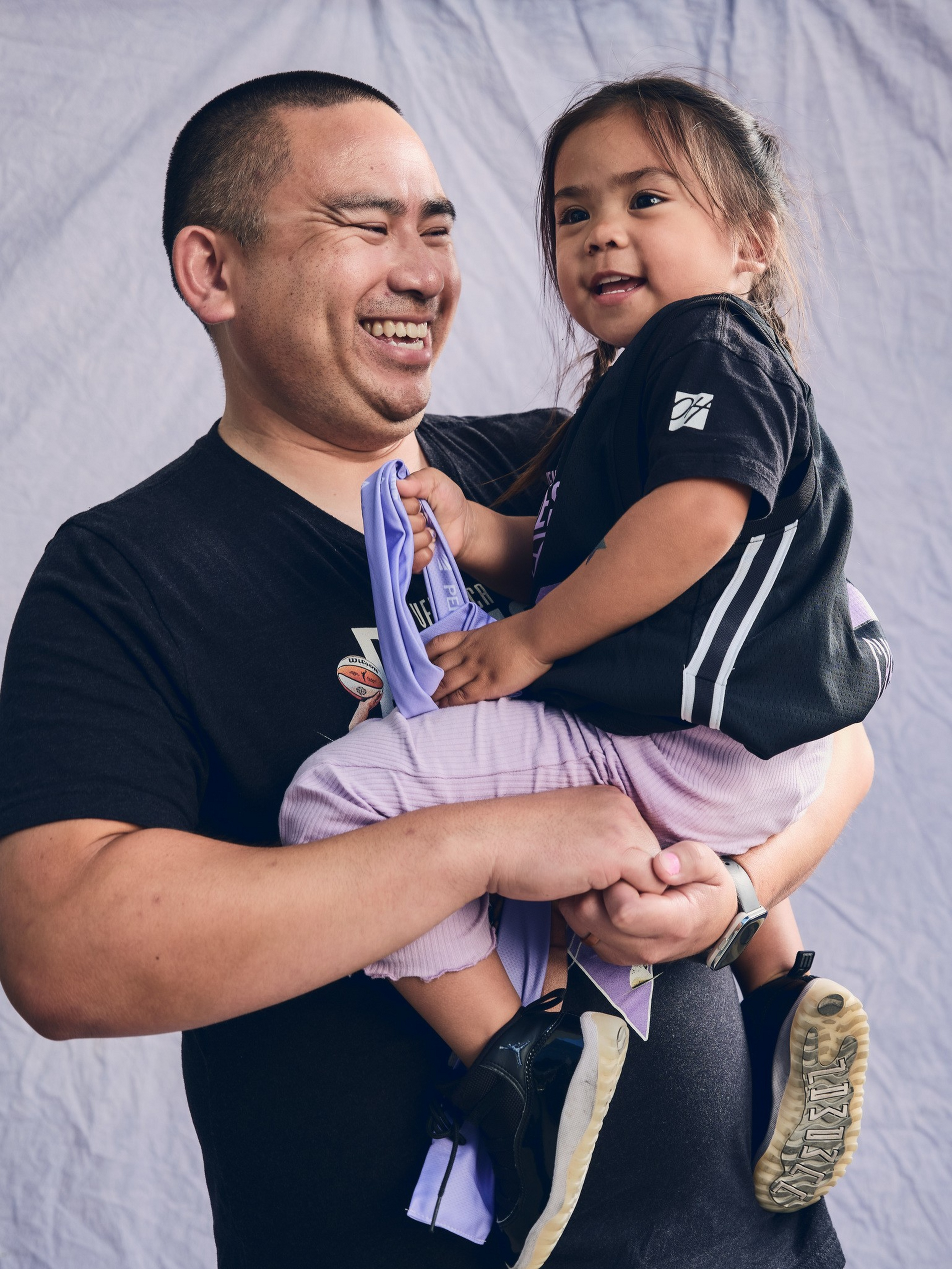 A man wearing a black t-shirt smiles while holding a young girl in a black shirt and light purple pants, who is also smiling and holding a purple cloth.