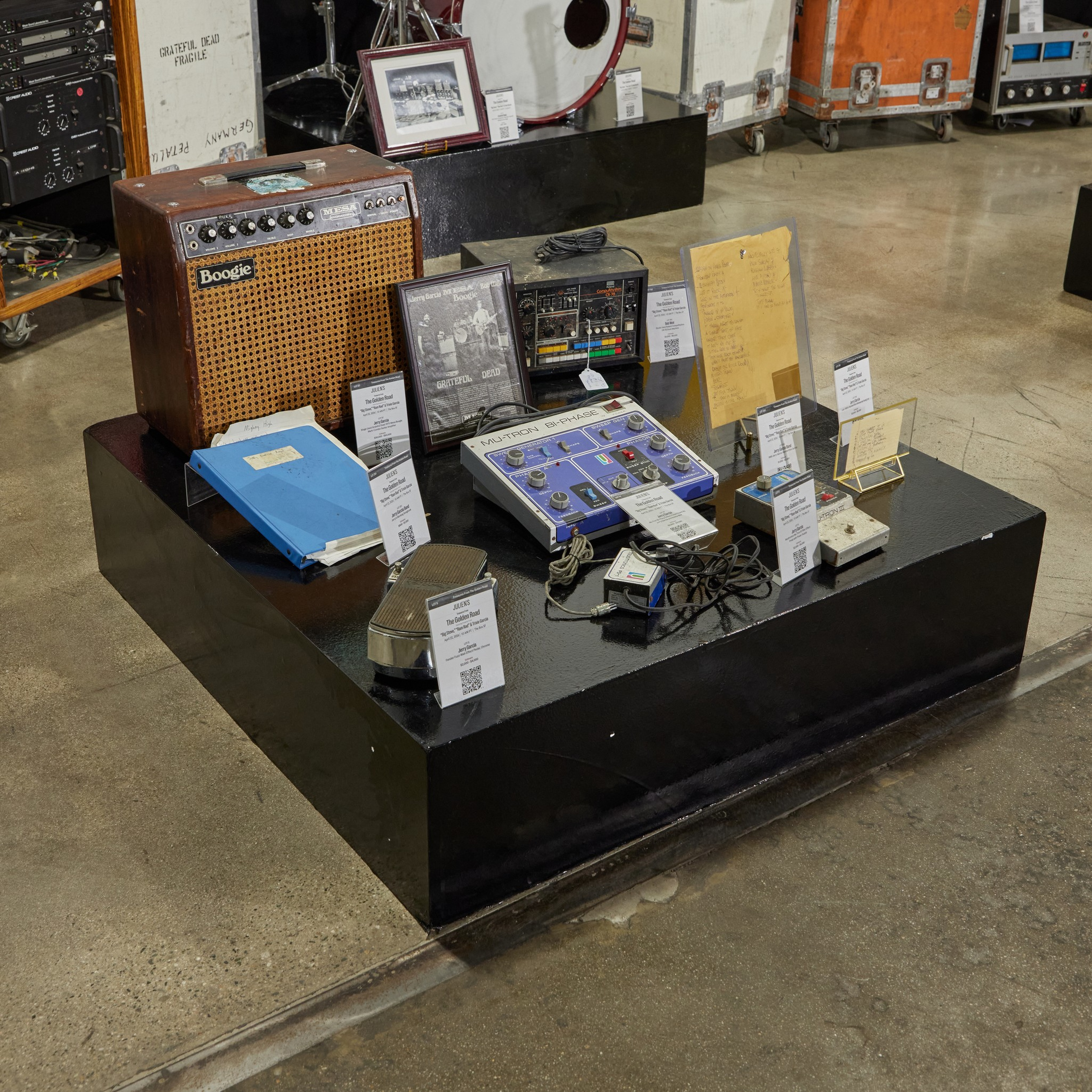 A display of vintage music equipment including a Boogie amplifier, effect pedals, a framed band photo, a blue binder, handwritten notes, and various cables on a black platform.