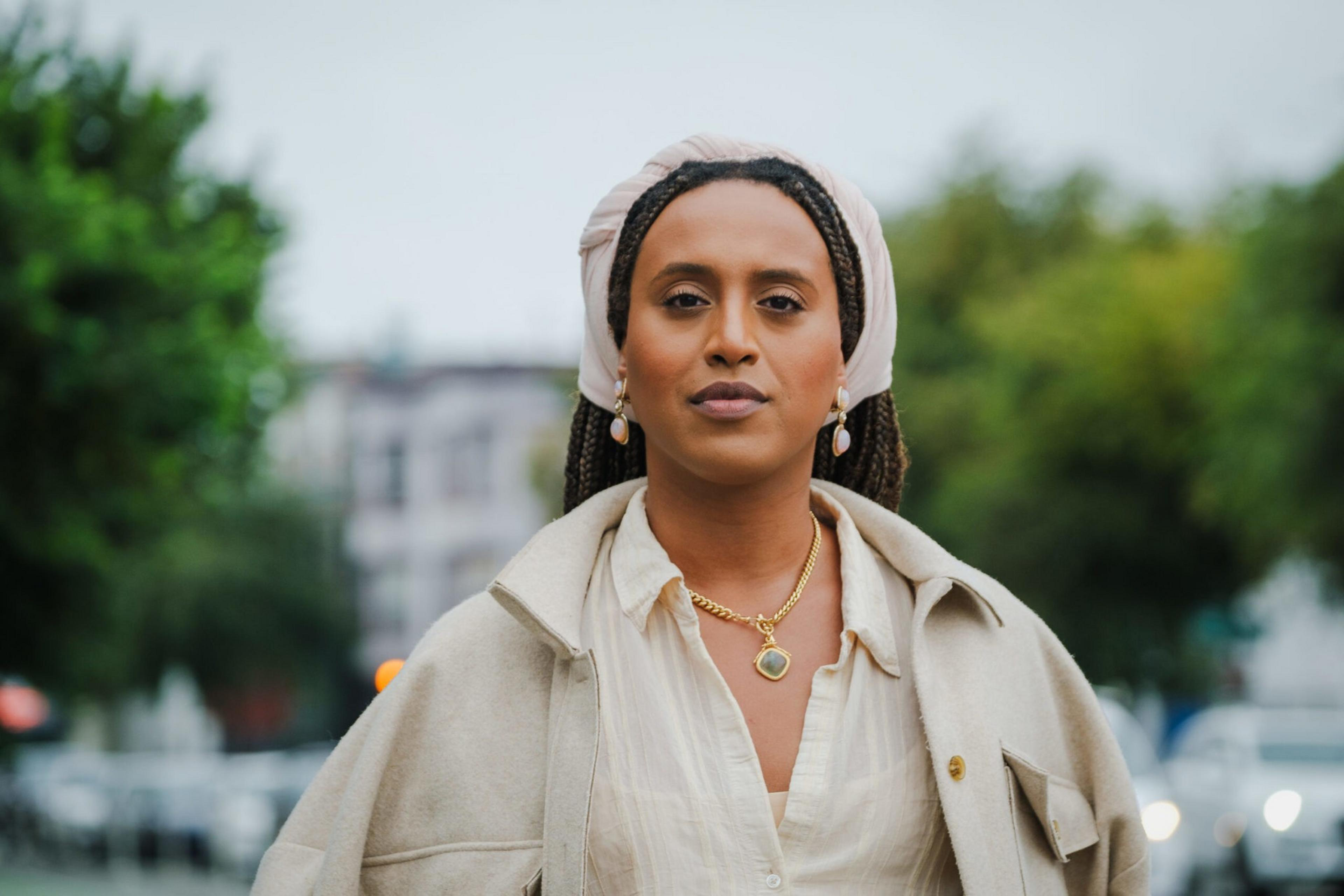 A confident woman with braided hair wears a beige headscarf, gold necklace, earrings, and a light beige coat over a white shirt against a blurred outdoor background.