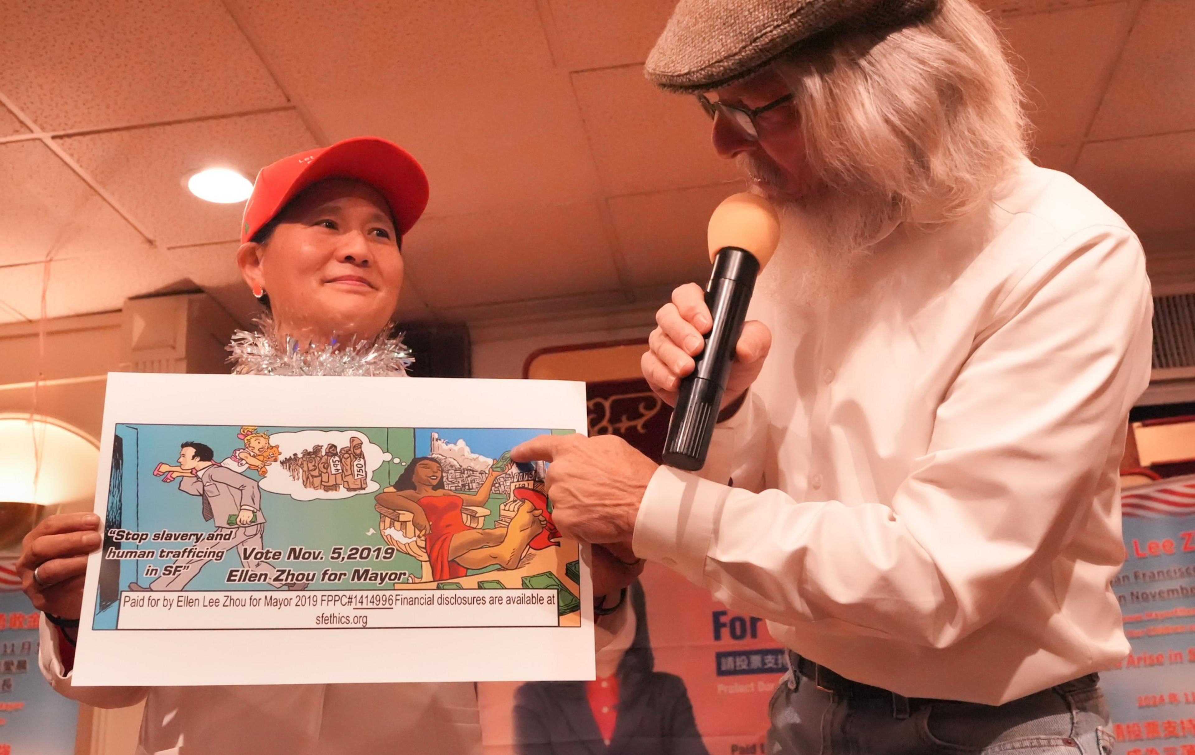 A person in a red cap is holding a colorful campaign sign beside a bearded man with a microphone pointing at the sign.