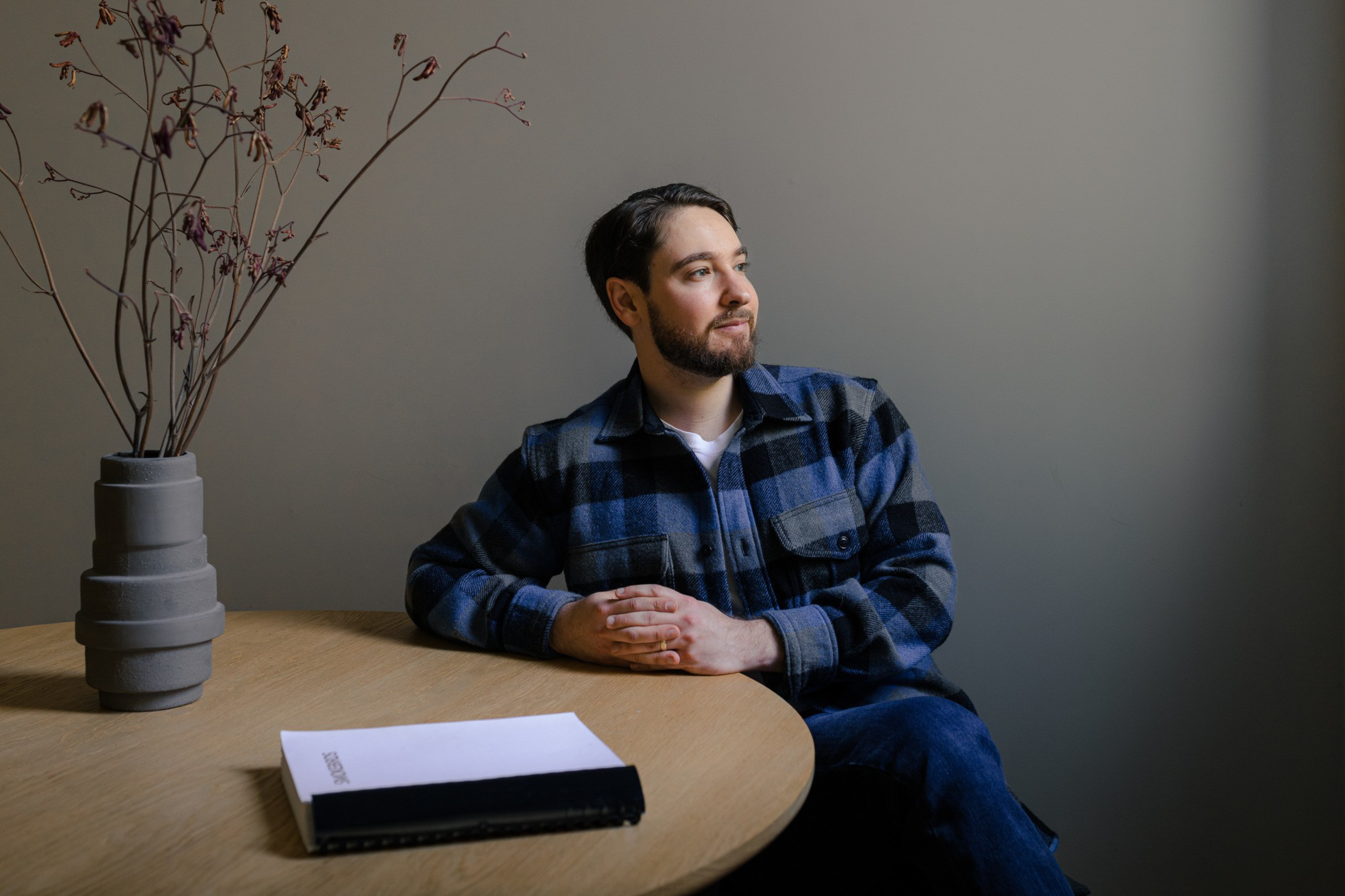A man in a checkered jacket sits at a round wooden table, gazing thoughtfully to the side. A vase with dried branches and a closed book are also on the table.