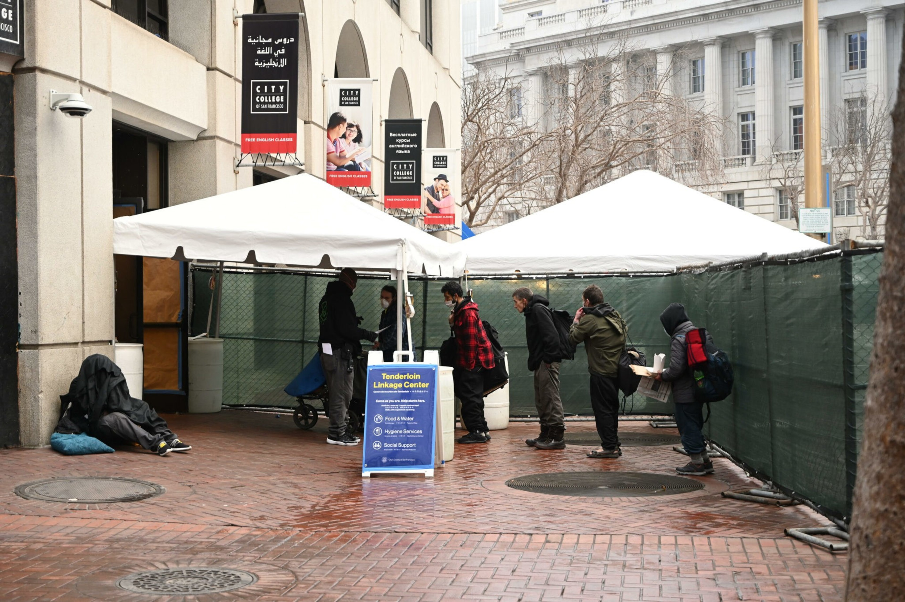 People stand in line outside a tent labeled "Tenderloin Linkage Center" offering food, water, hygiene, and social support services.