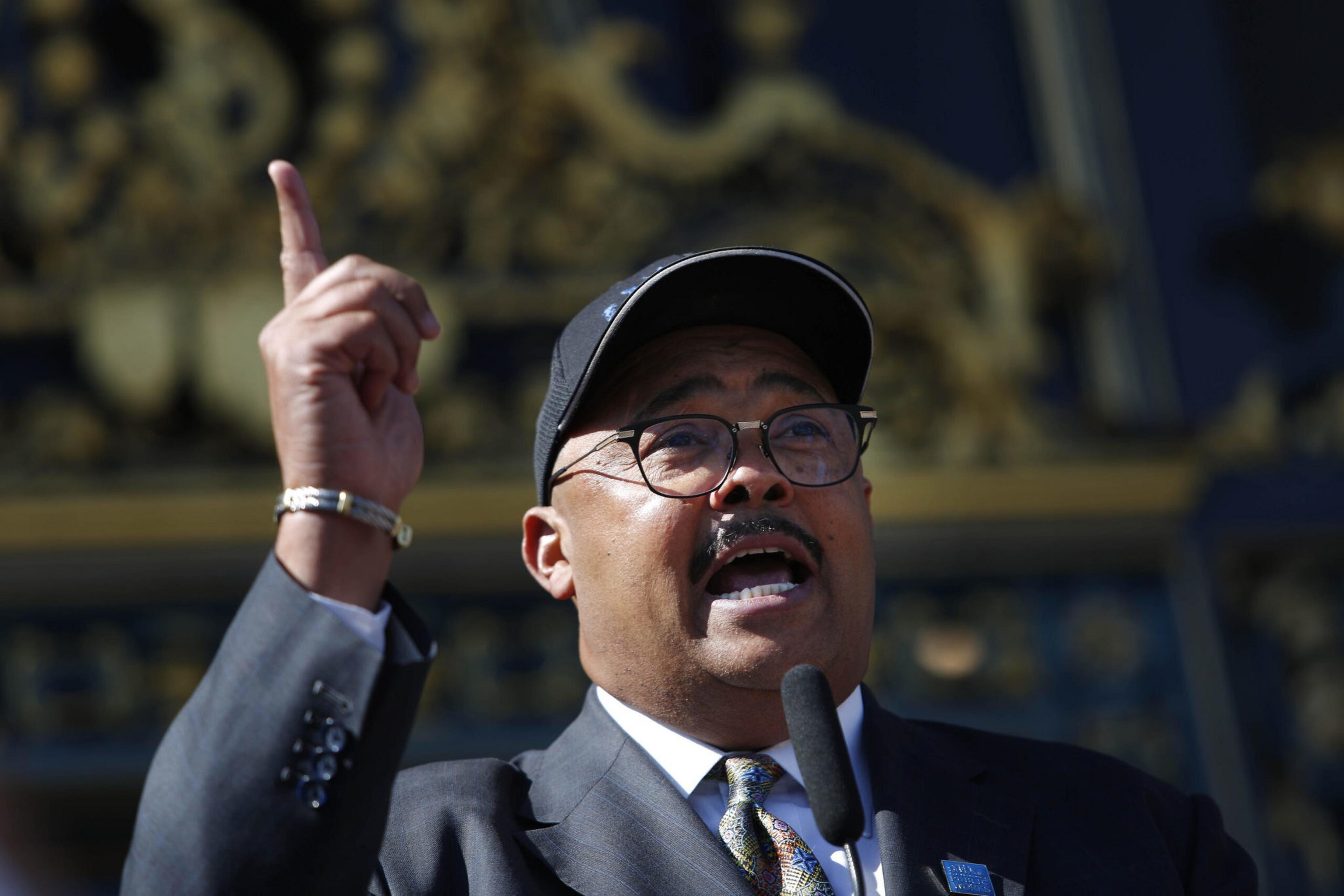 A man stands in front of a microphone while speaking and holding his finger in the air.
