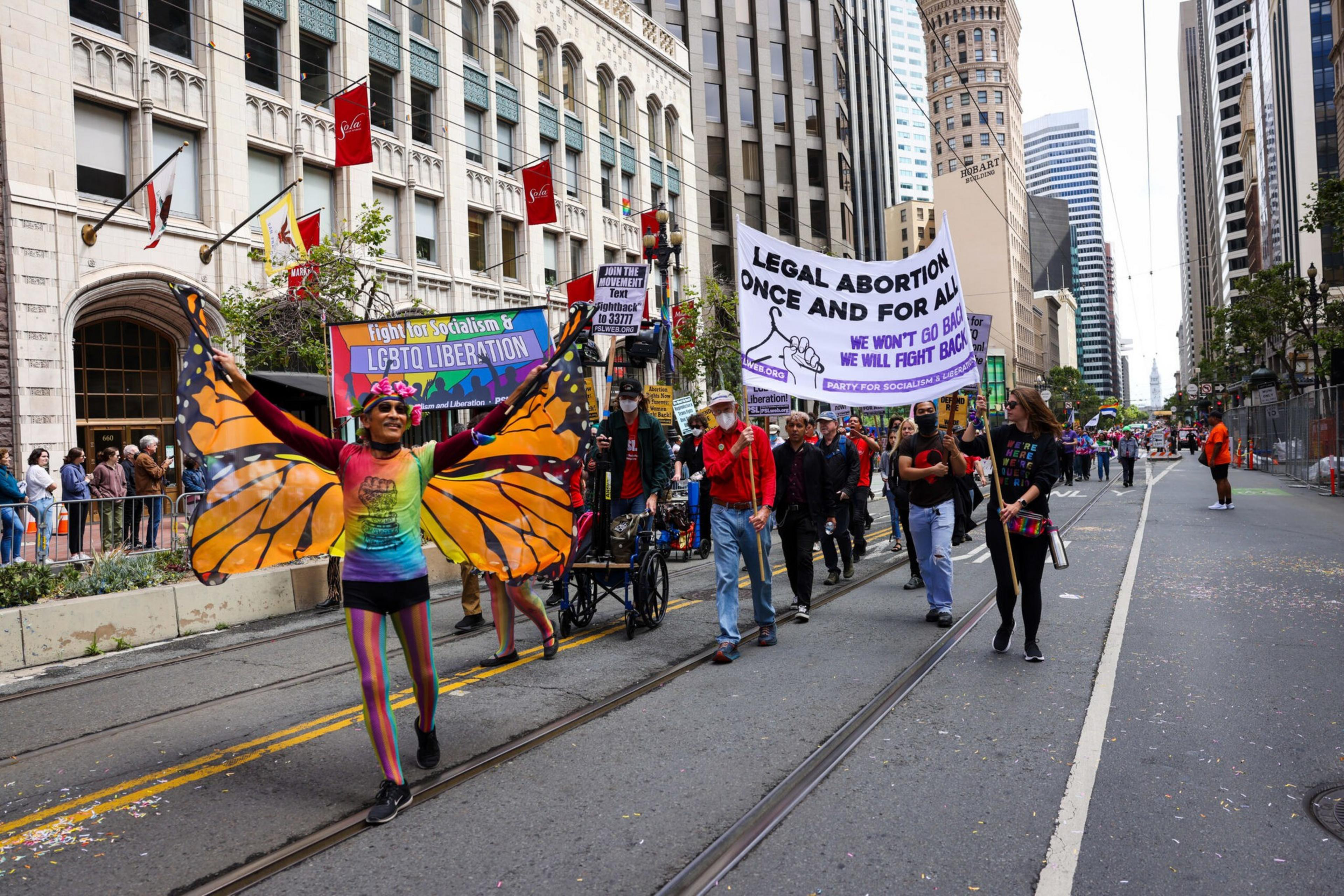 A person wearing large butterfly wings and rainbow clothes leads a march holding a sign for LGBTQ liberation and legal abortion rights in an urban street.