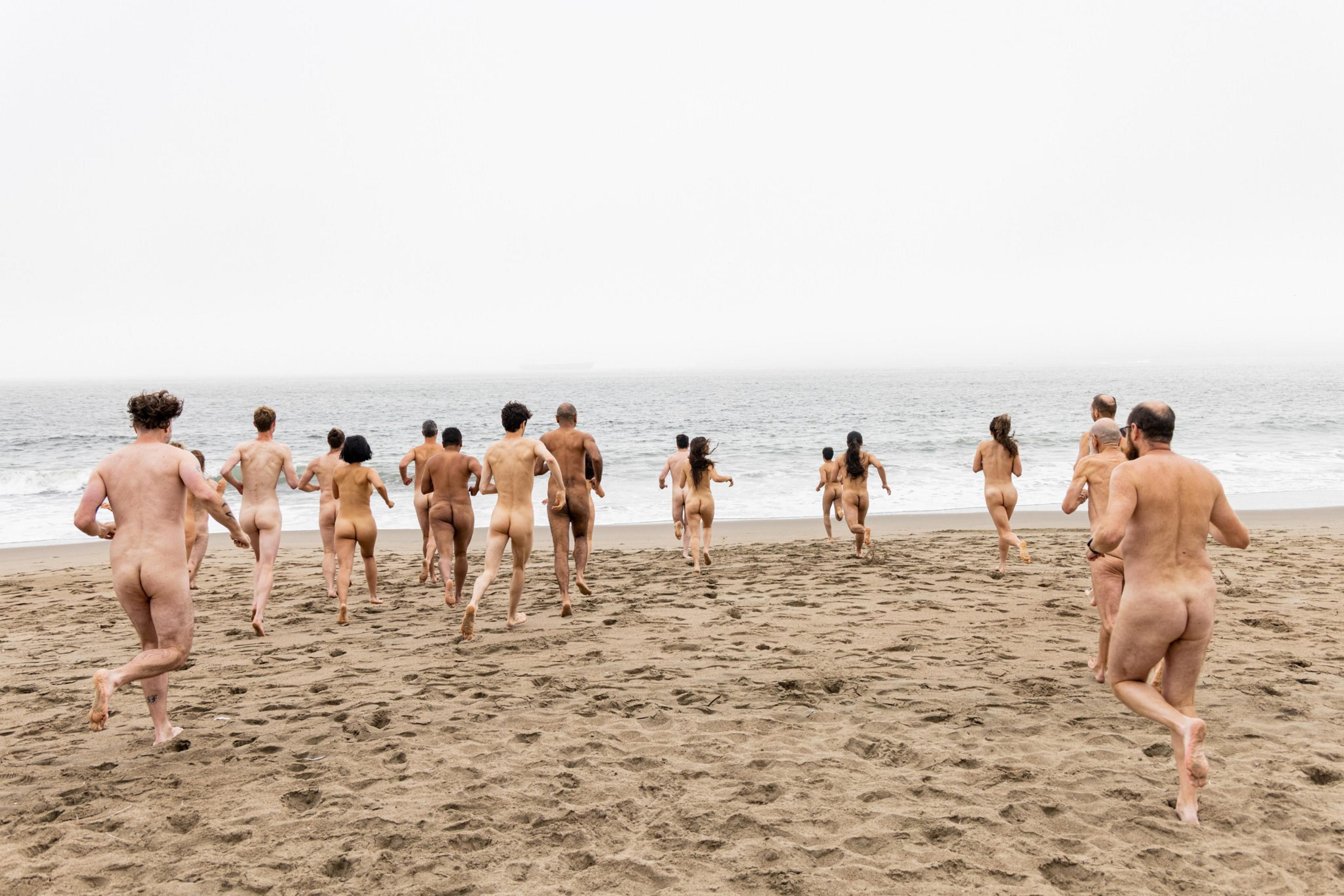A group of naked people are seen running towards the ocean on a sandy beach, under an overcast sky with mist in the distance.