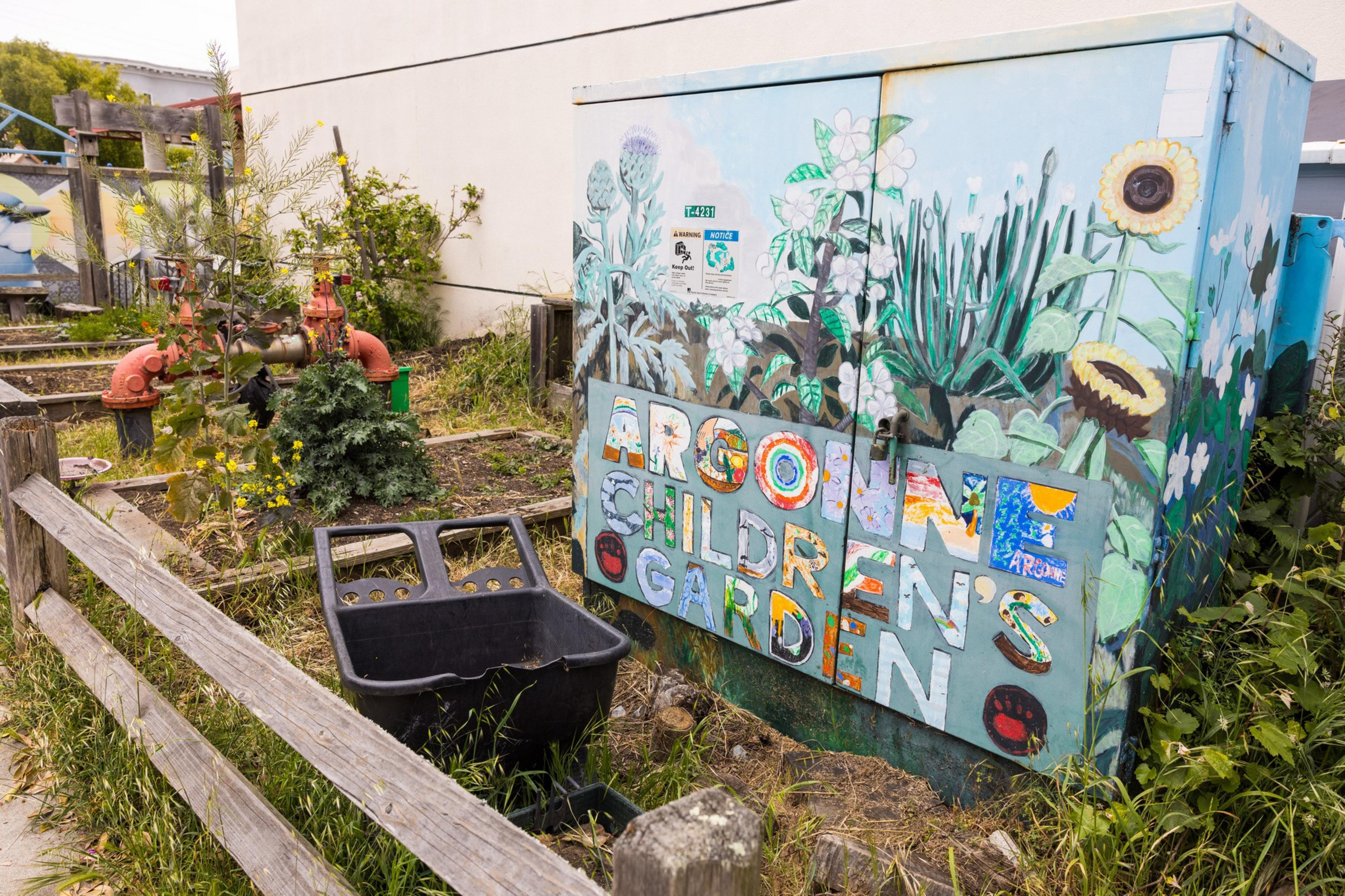 The image shows a colorful utility box in a garden area. It is painted with flowers and the words &quot;Argonne Children's Garden.&quot; There's a wheelbarrow and a wooden fence nearby.