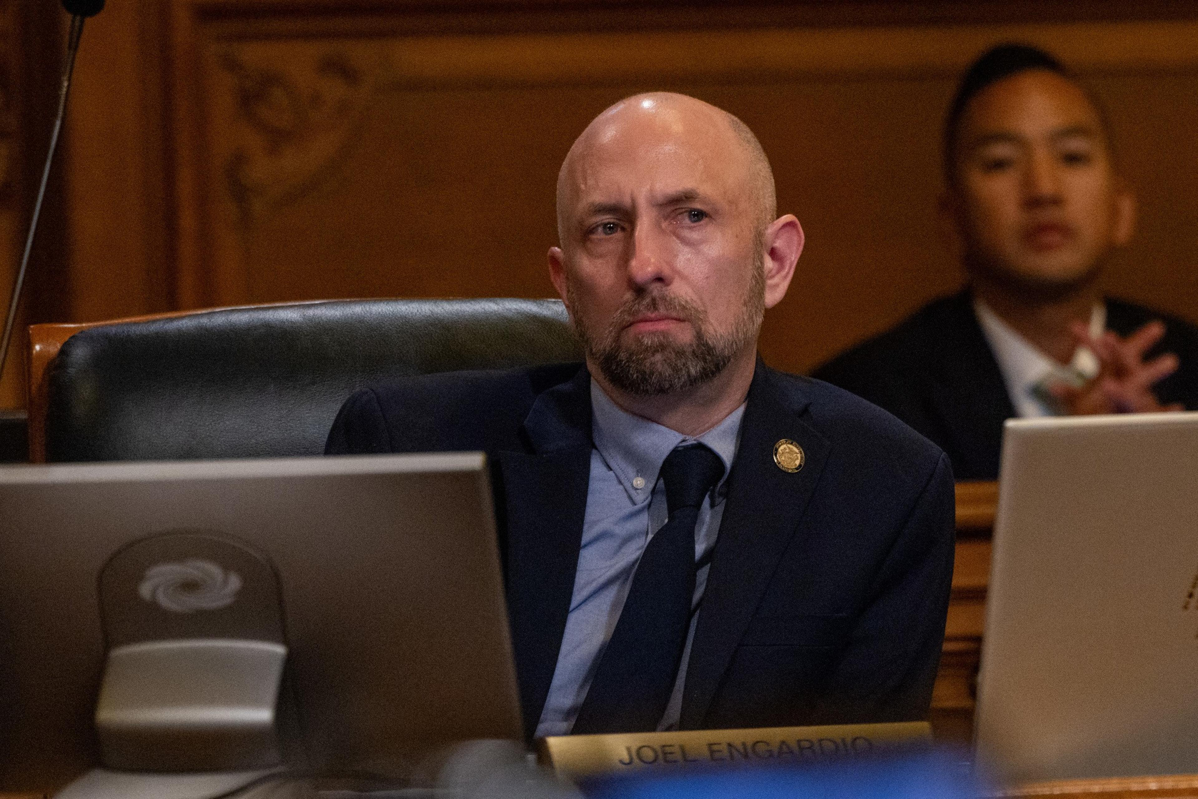 A bald man with a beard wearing a suit and tie sits at a desk, looking to the side thoughtfully, with another person blurred in the background.
