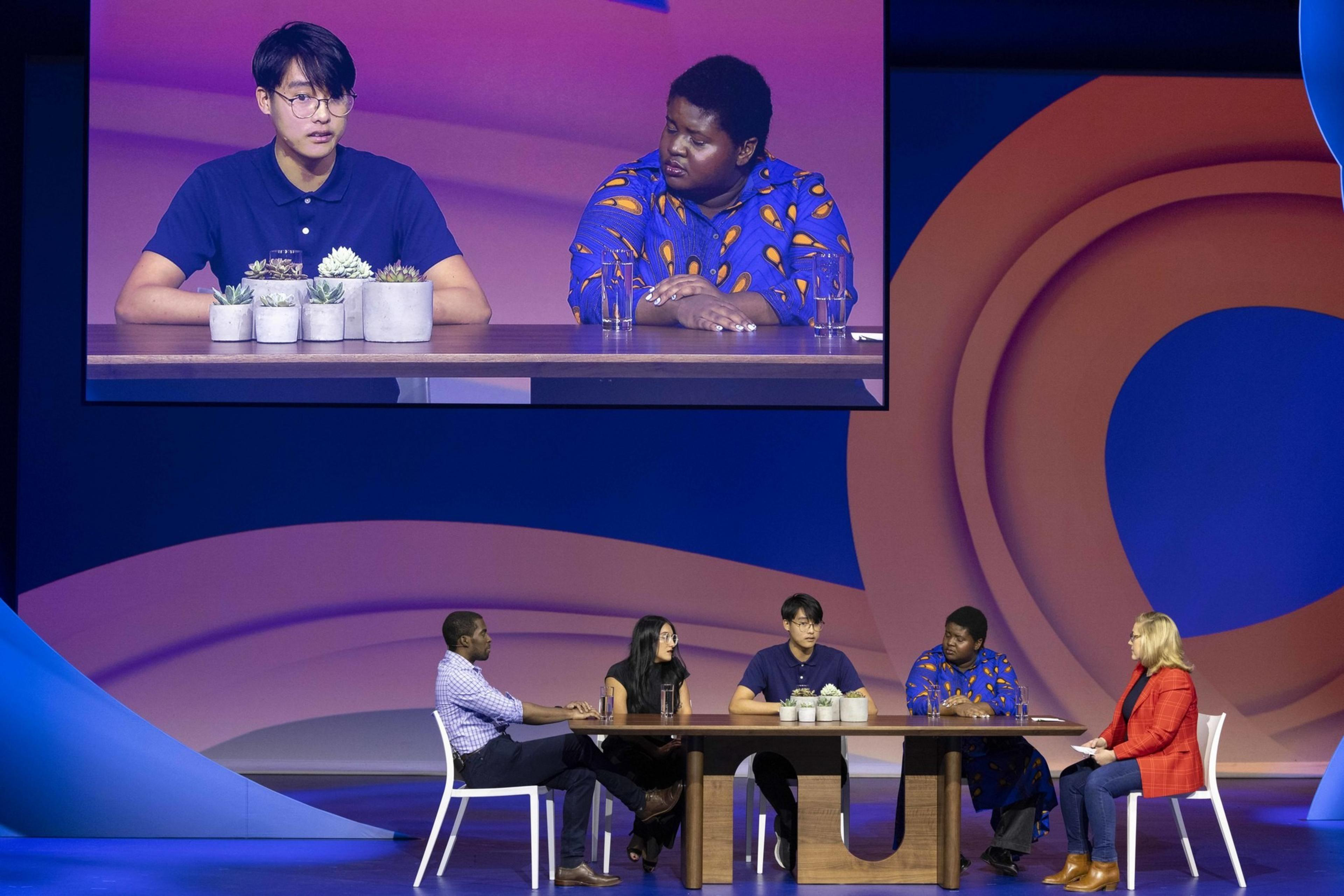 Five people sit at a table on stage, engaged in a discussion. A large screen above displays a close-up of two of them, with potted plants in view.