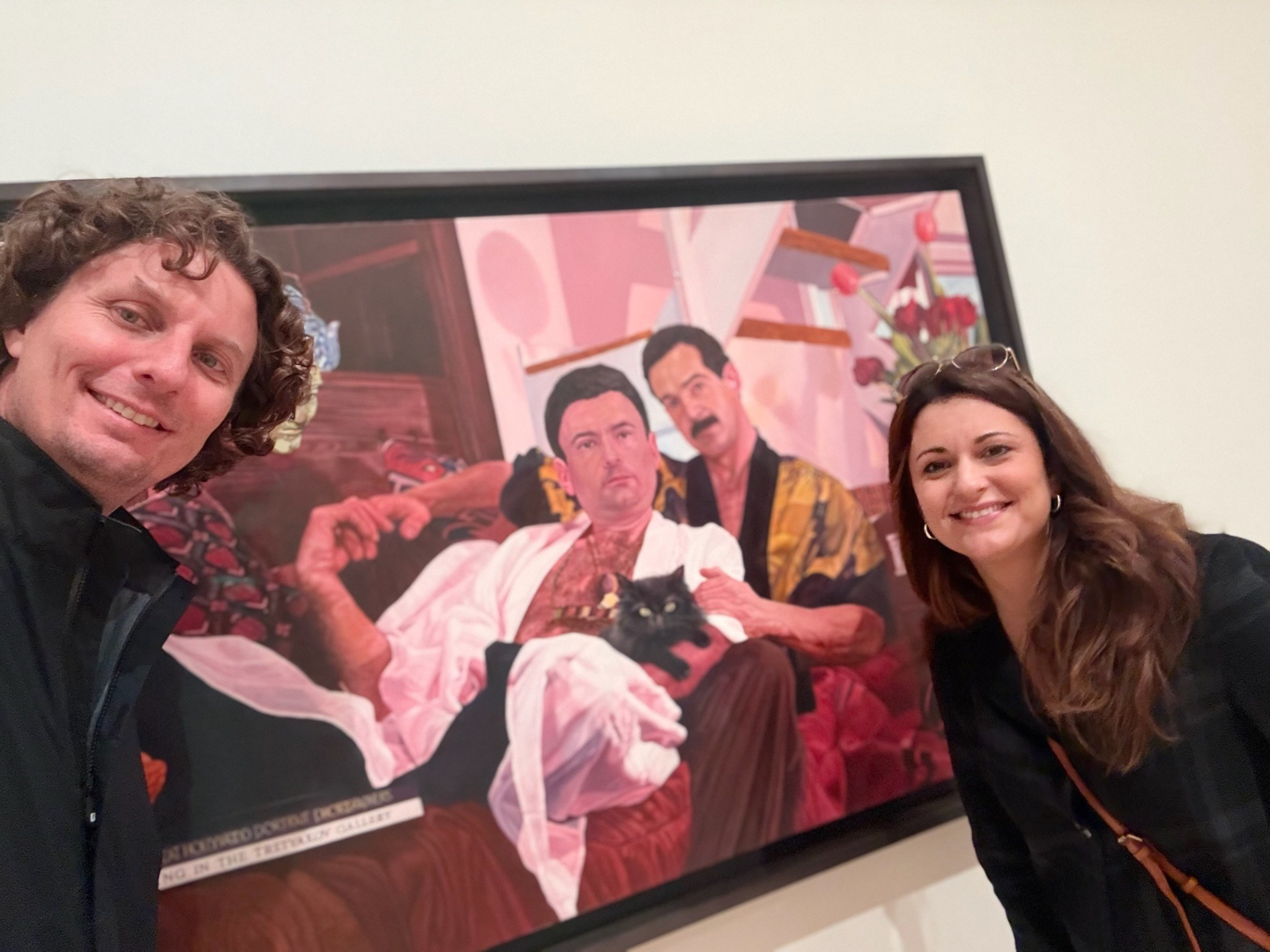 A man and a woman smile beside a framed painting of two men sitting on ...