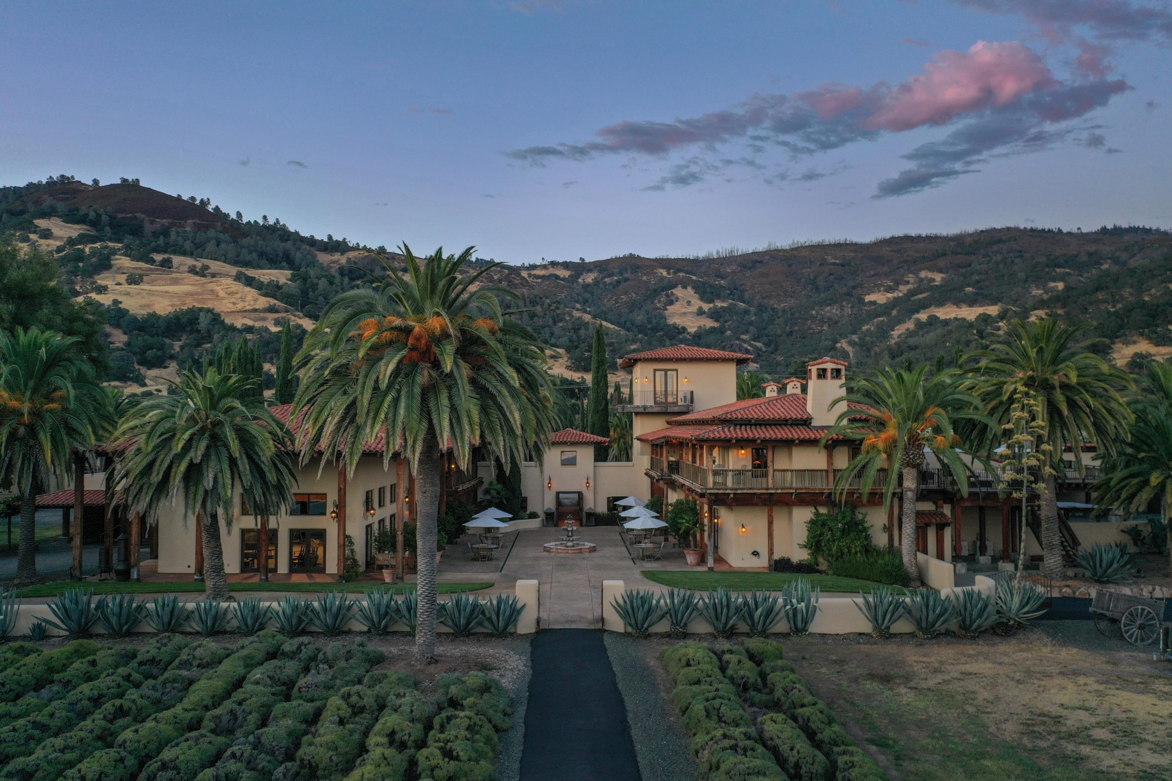 A large Mediterranean-style house with red tile roofs is surrounded by palm trees and shrubbery, set against rolling hills under a twilight sky.