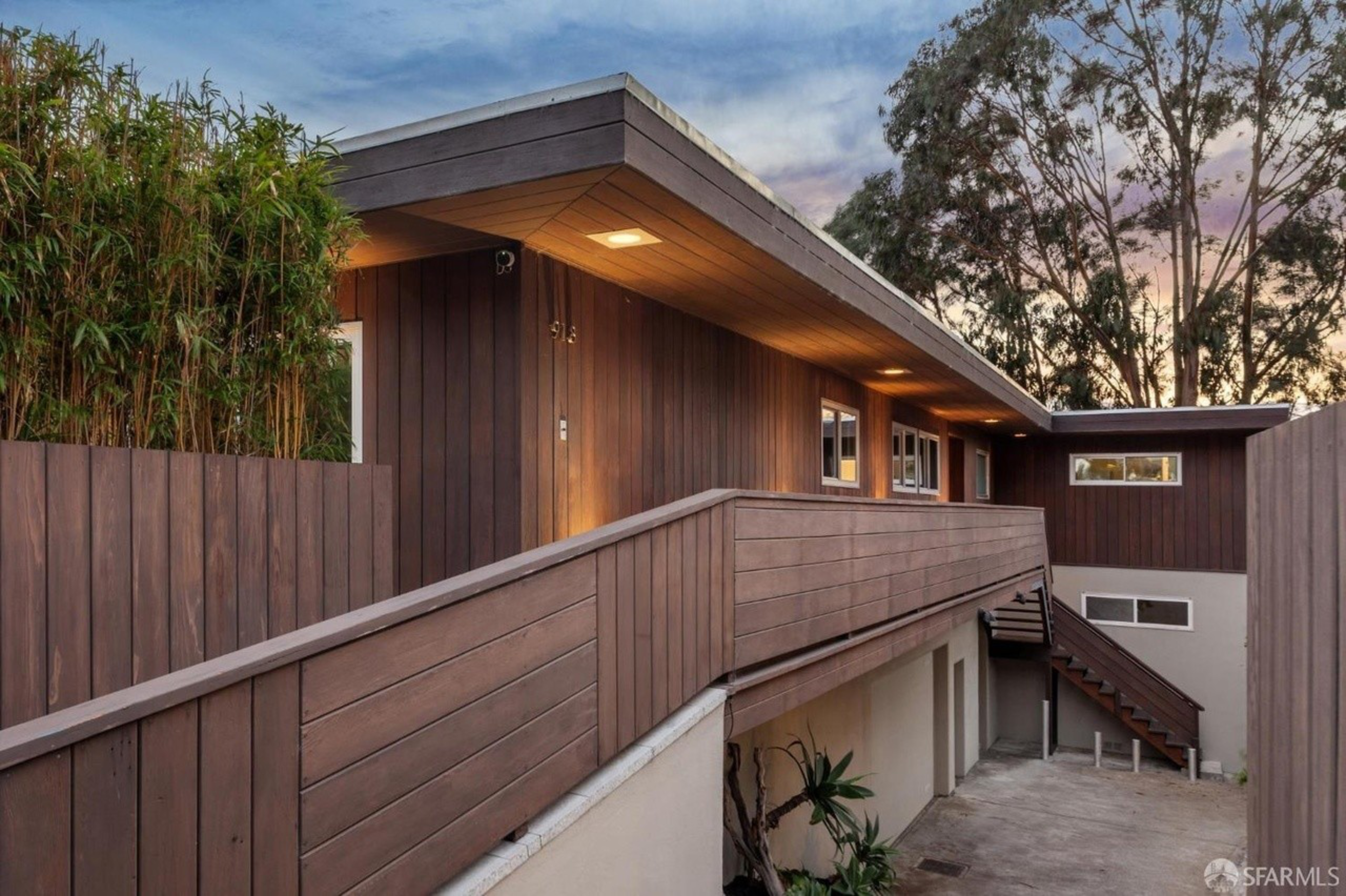 A modern house with dark wooden paneling, an elevated walkway with railings, stairs, and surrounding greenery under a dusk sky.