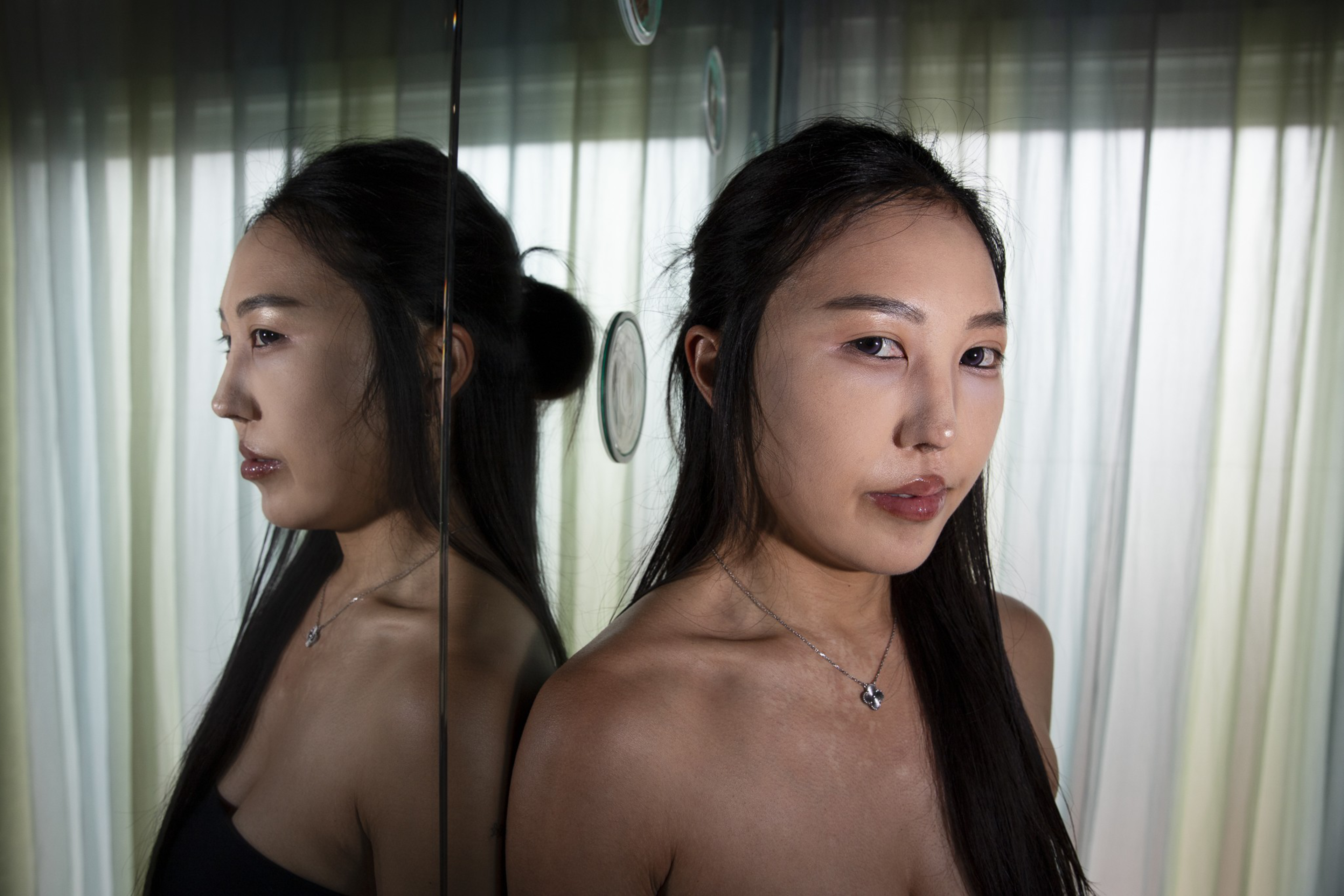 A woman with long dark hair and a necklace stands against a reflective surface, showing her front and side profile under soft lighting.