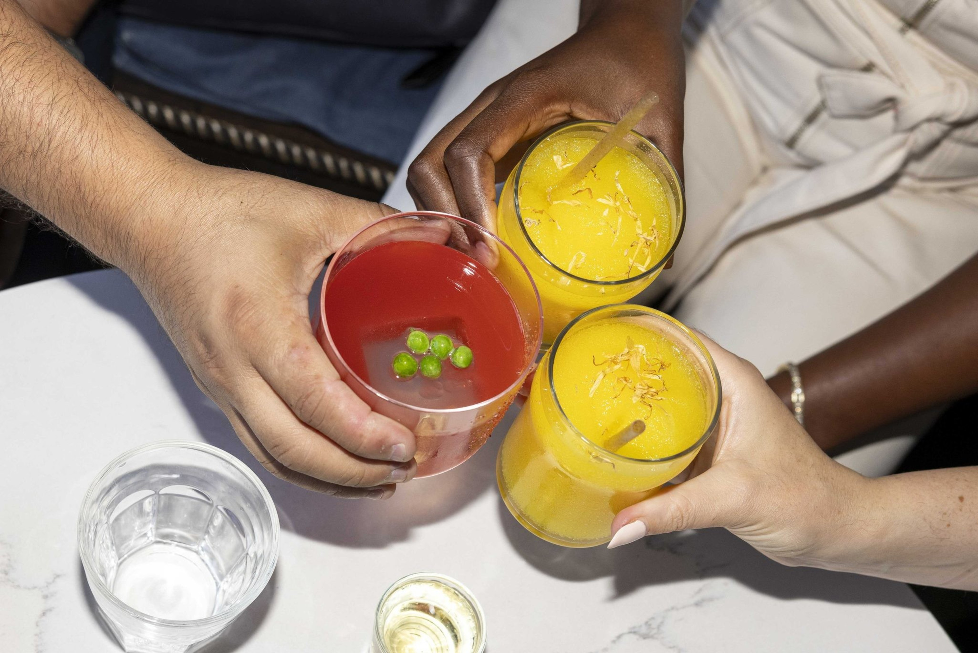 Three hands hold colorful drinks, toasting above a table with fries, ketchup, mustard, a glass of water, and two small shot glasses.