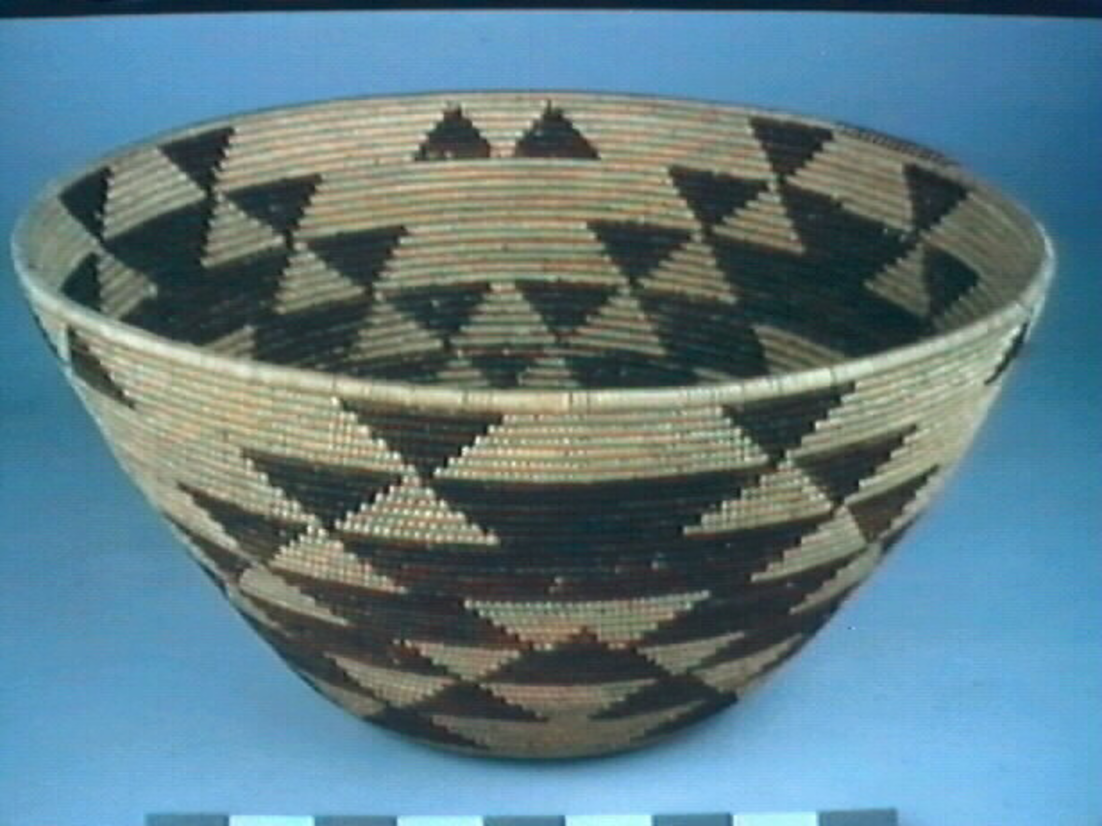 A woven basket with a wide opening and narrow base, decorated with repeated black and natural-colored triangular geometric patterns.