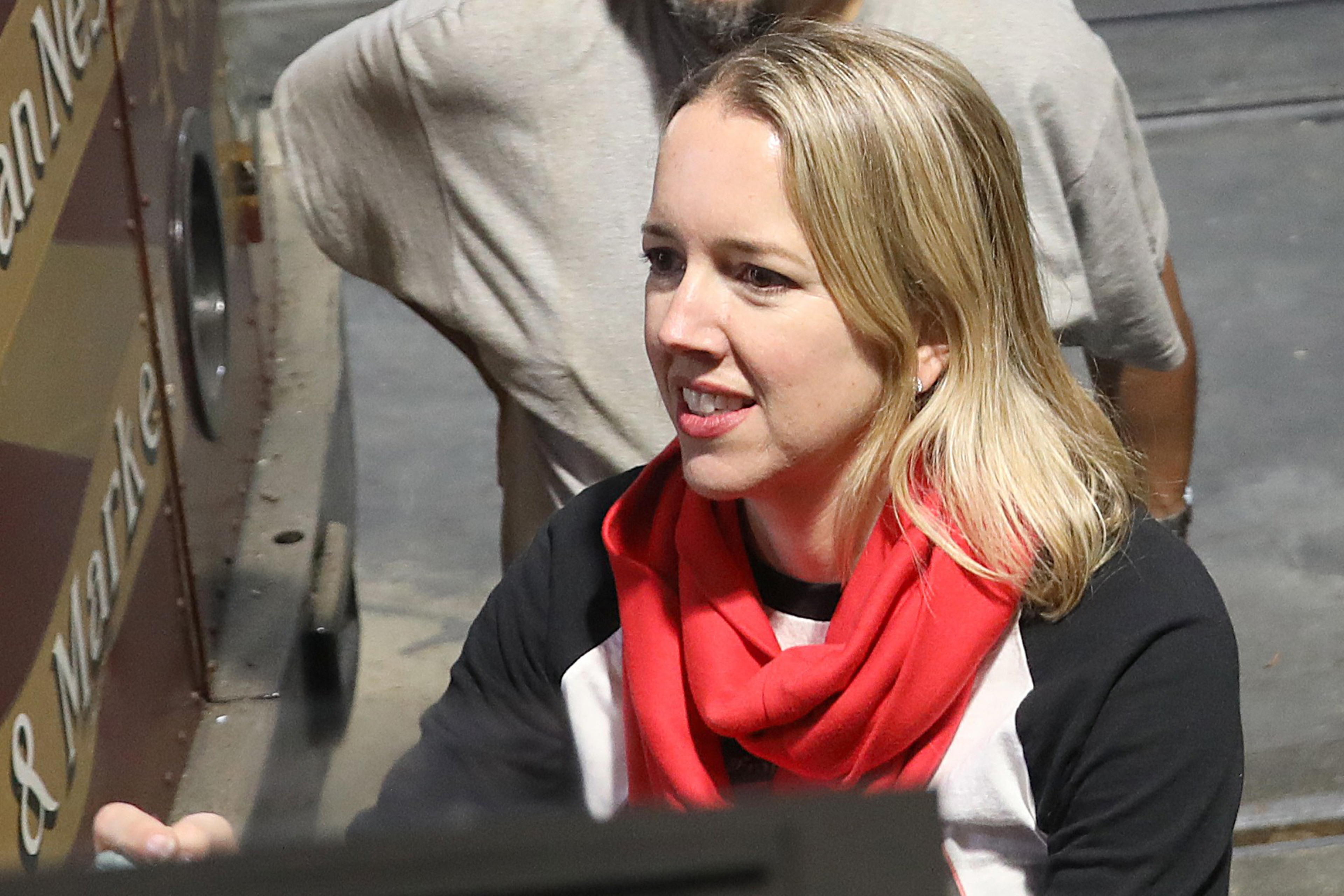 A blonde woman wearing a black and white top with a red scarf smiles while interacting with someone outside the frame, with a man standing behind her.
