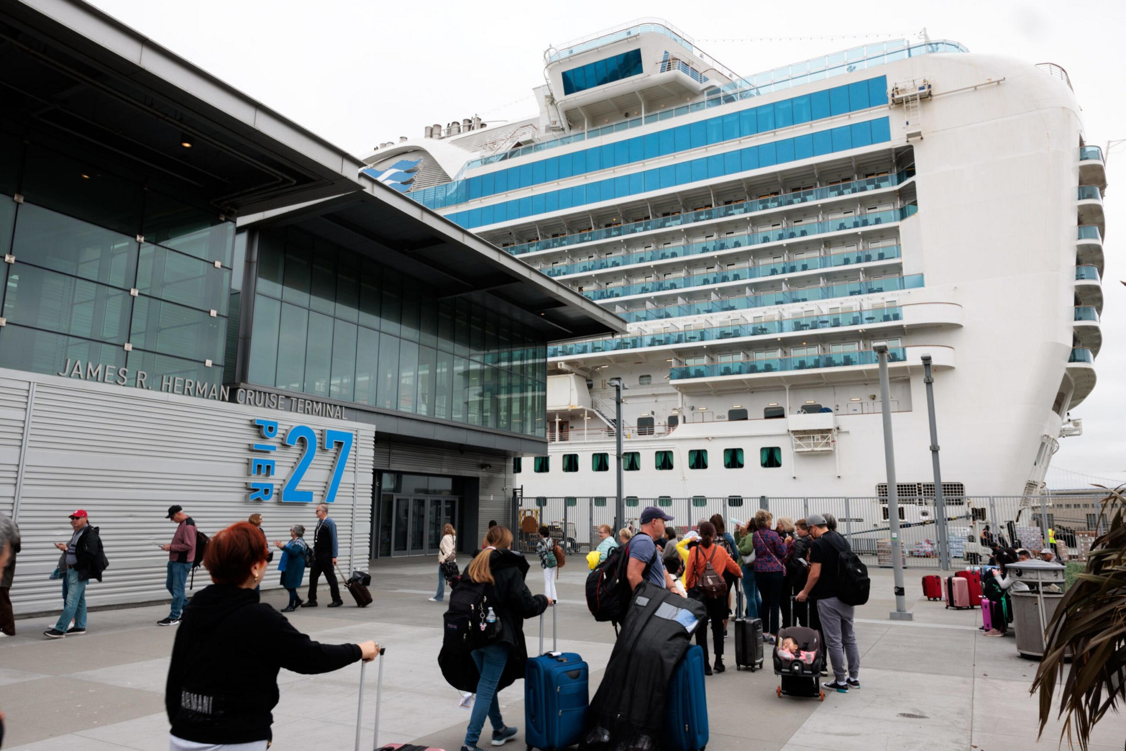 Passengers with luggage wait in line outside the James R. Herman Cruise Terminal at Pier 27 beside a large docked cruise ship.