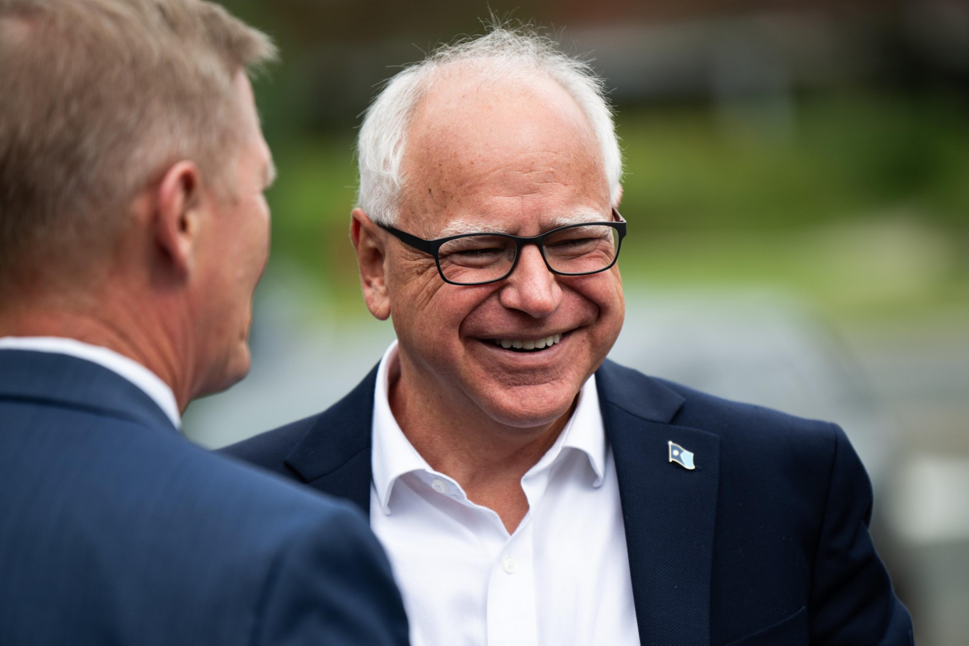 Two men in suits are outdoors, one with white hair and glasses smiling warmly, engaging in conversation, with a blurred green background.