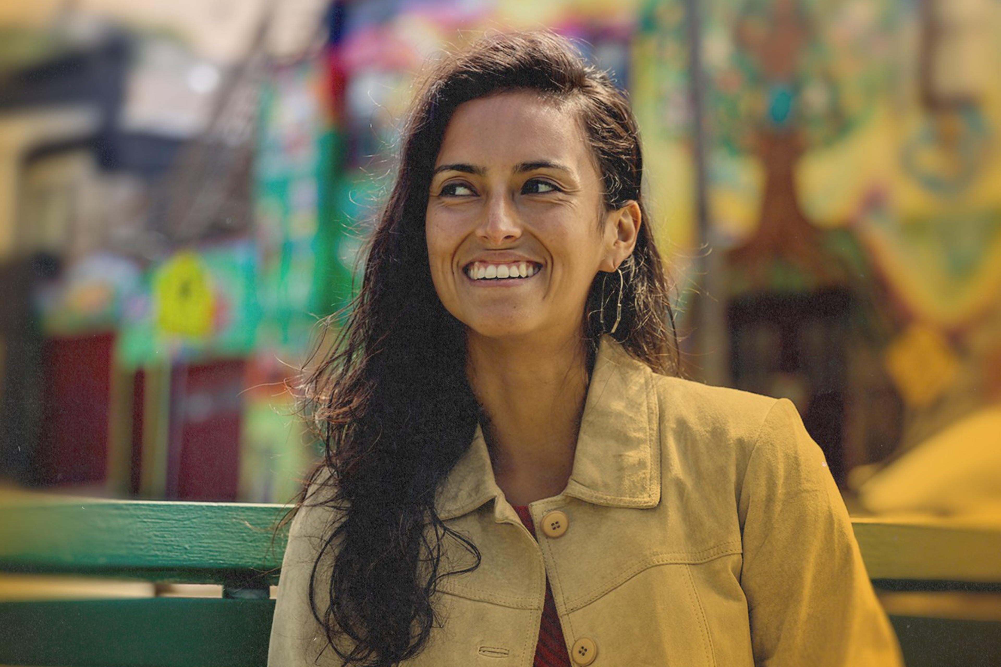 A woman with long dark hair is smiling brightly while sitting on a green bench. She wears a tan jacket, and behind her is a colorful, blurred background.