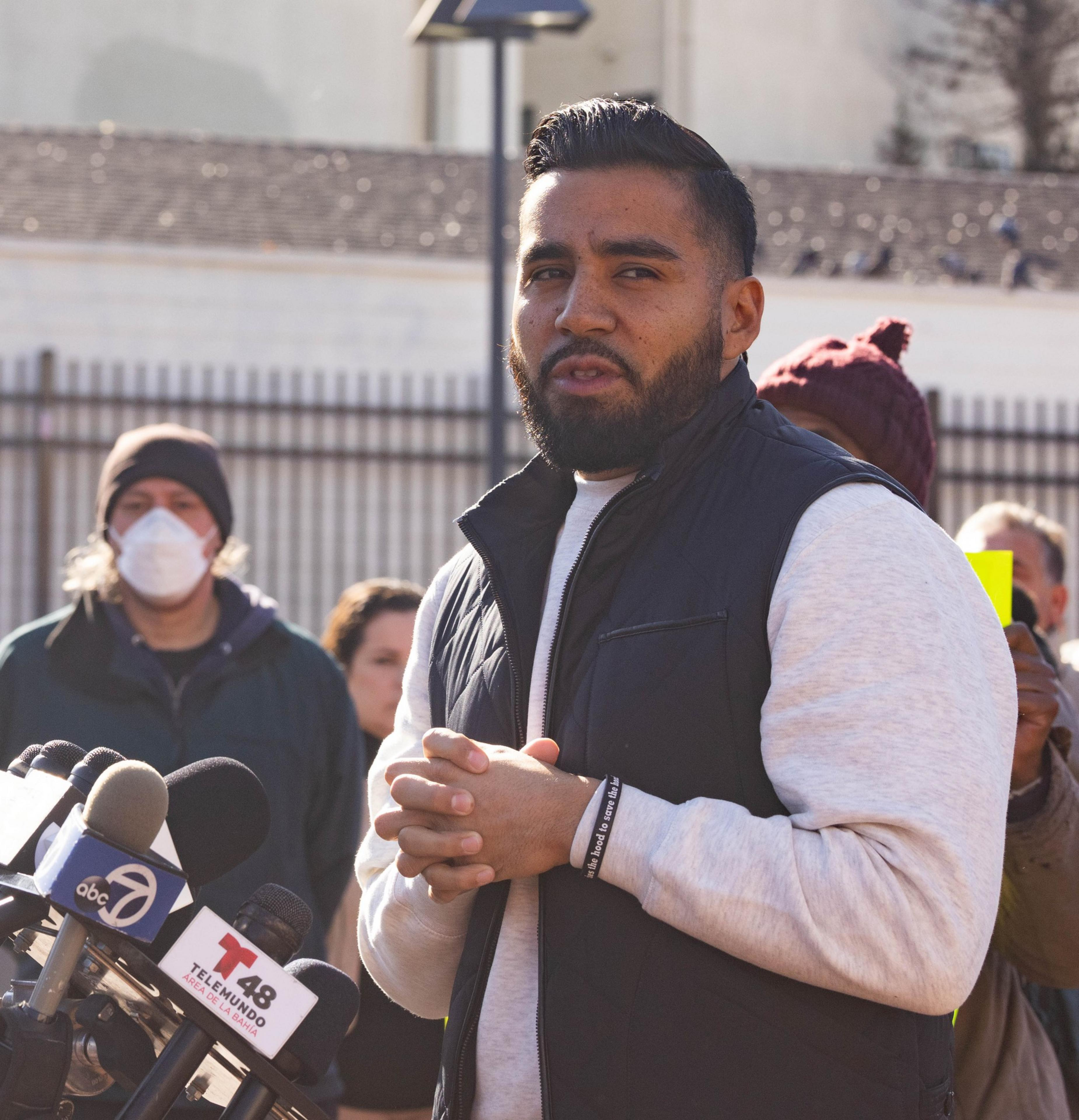 Jon Jacobo is speaking at a press conference with microphones in front, and onlookers behind him.