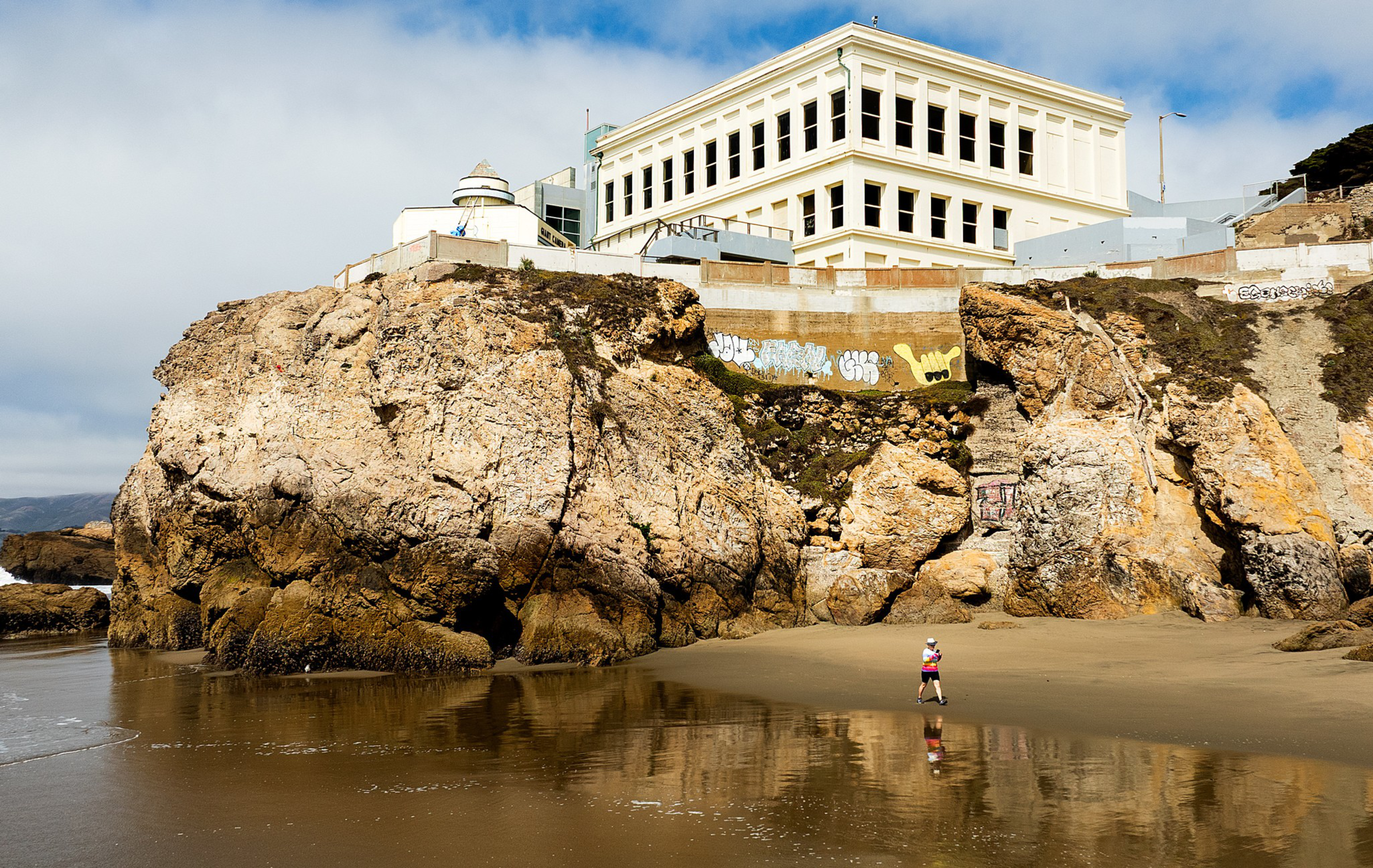 A large rocky cliff with graffiti supports a white building by the beach, where a person in a pink jacket walks on wet sand near calm water.