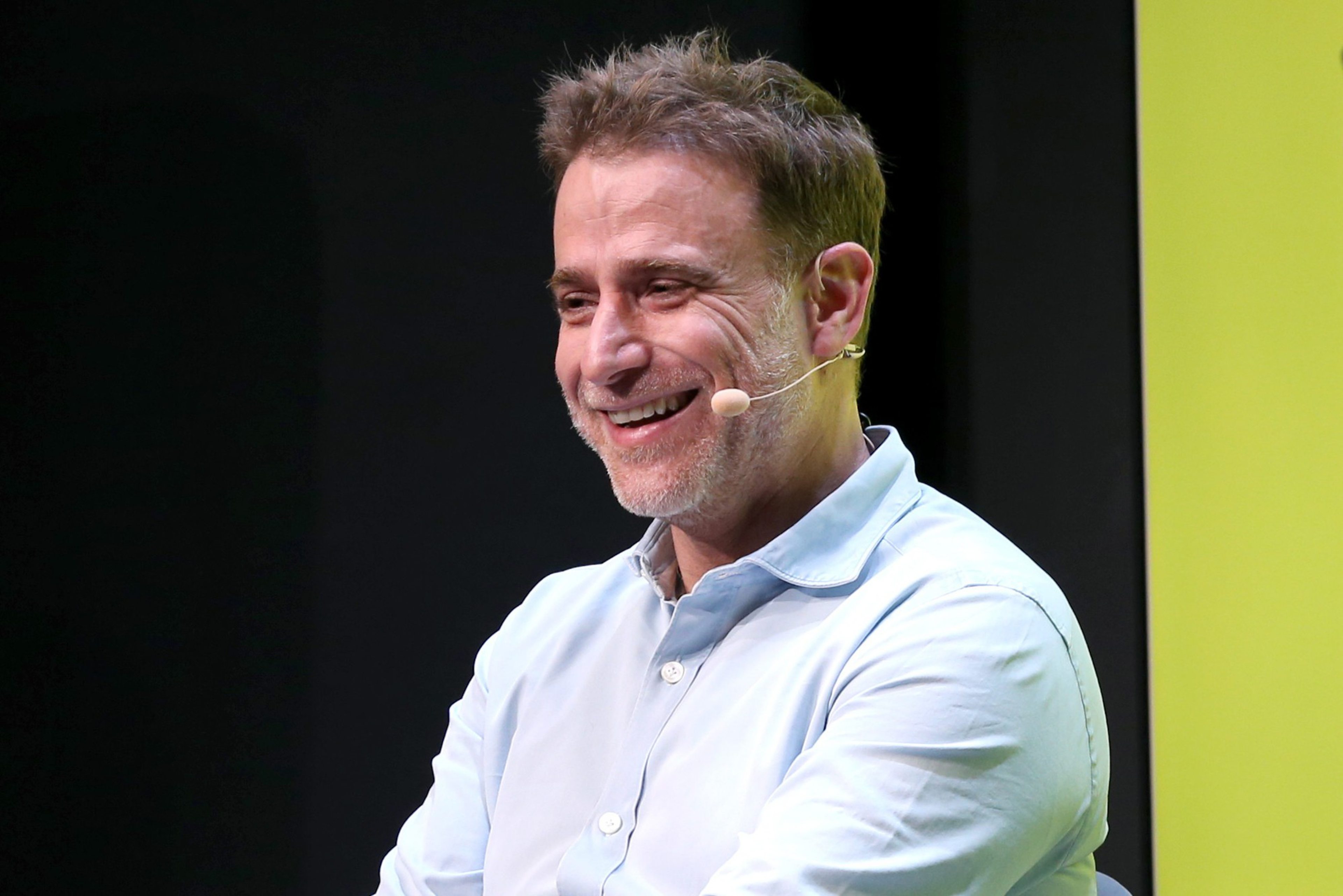 A man with short brown hair and a light beard smiles while wearing a light blue shirt and a headset microphone against a dark background.