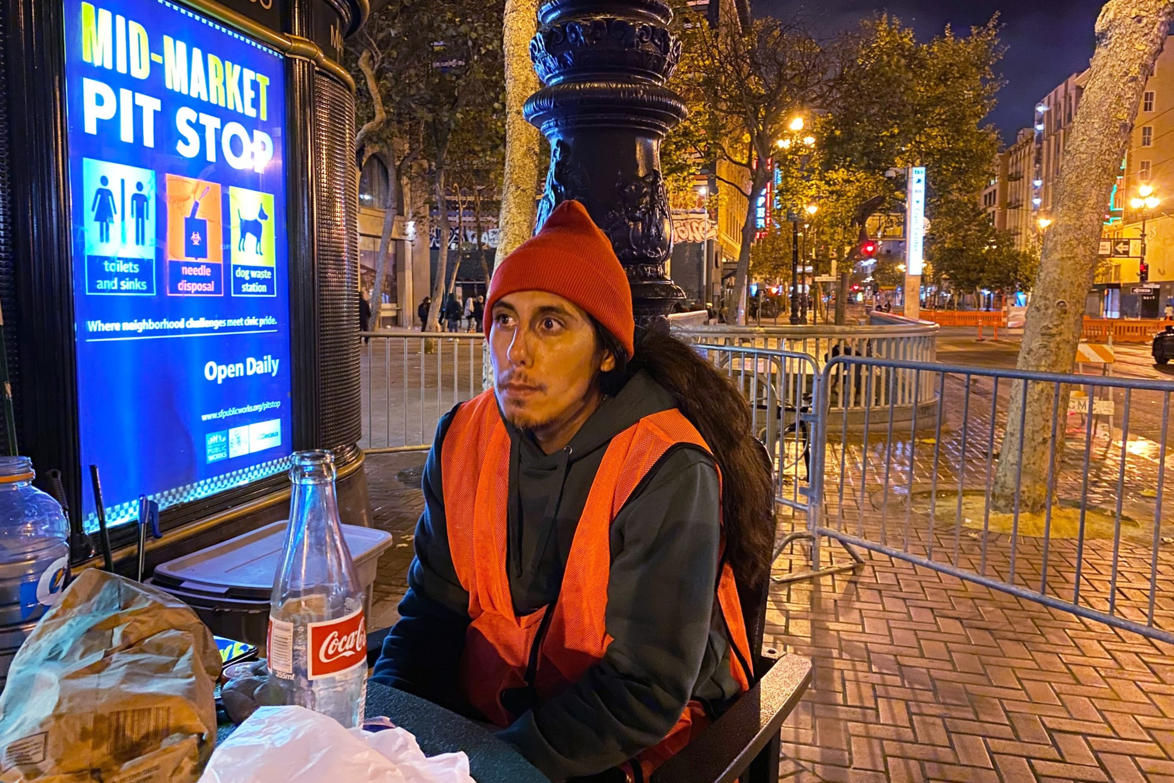 A person sits at night by a lit-up &quot;Pit Stop&quot; sign; street lights and trees in the background.