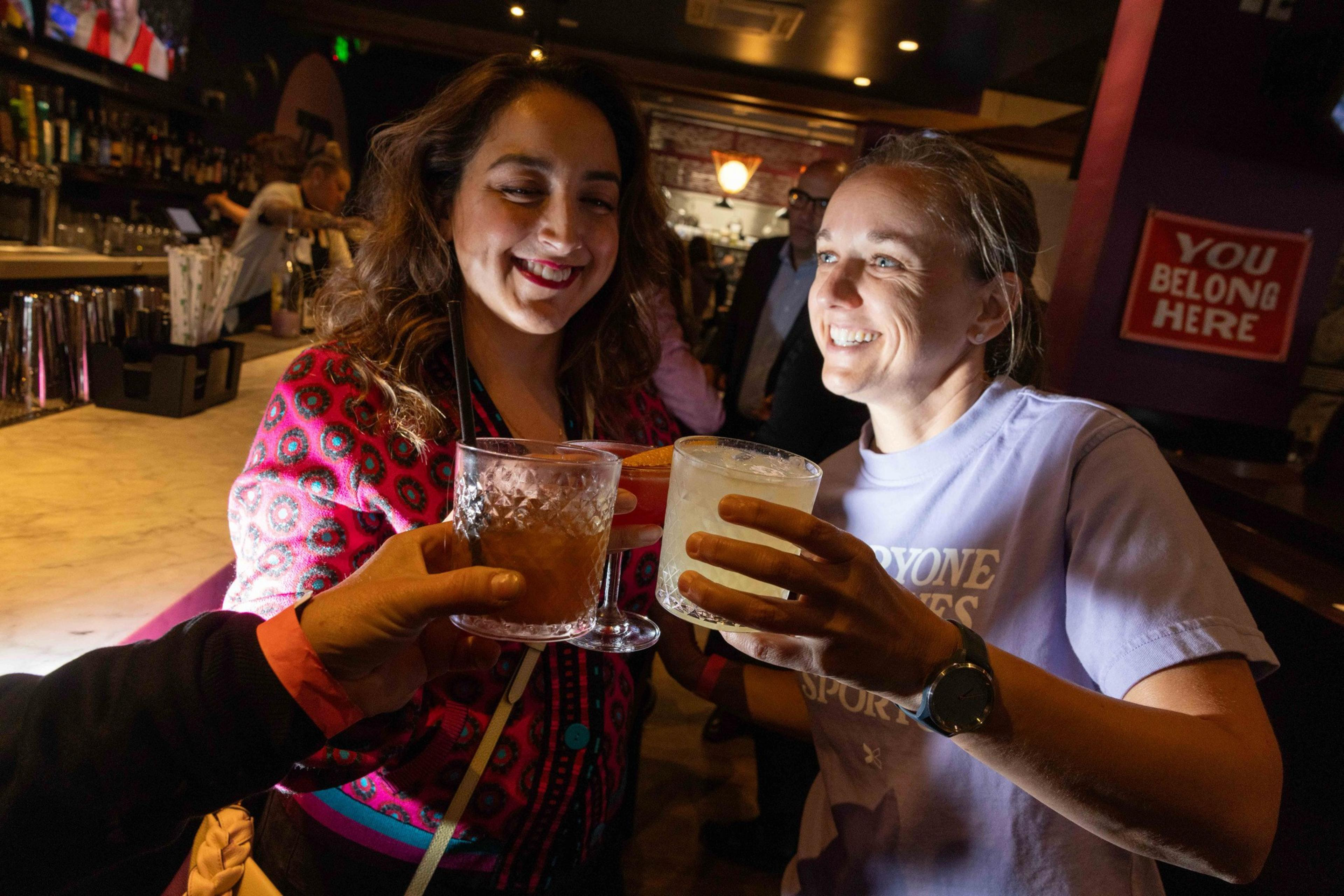Two women are smiling and clinking glasses with someone at a bar, where a sign reads &quot;You Belong Here.&quot; The atmosphere is lively and welcoming.