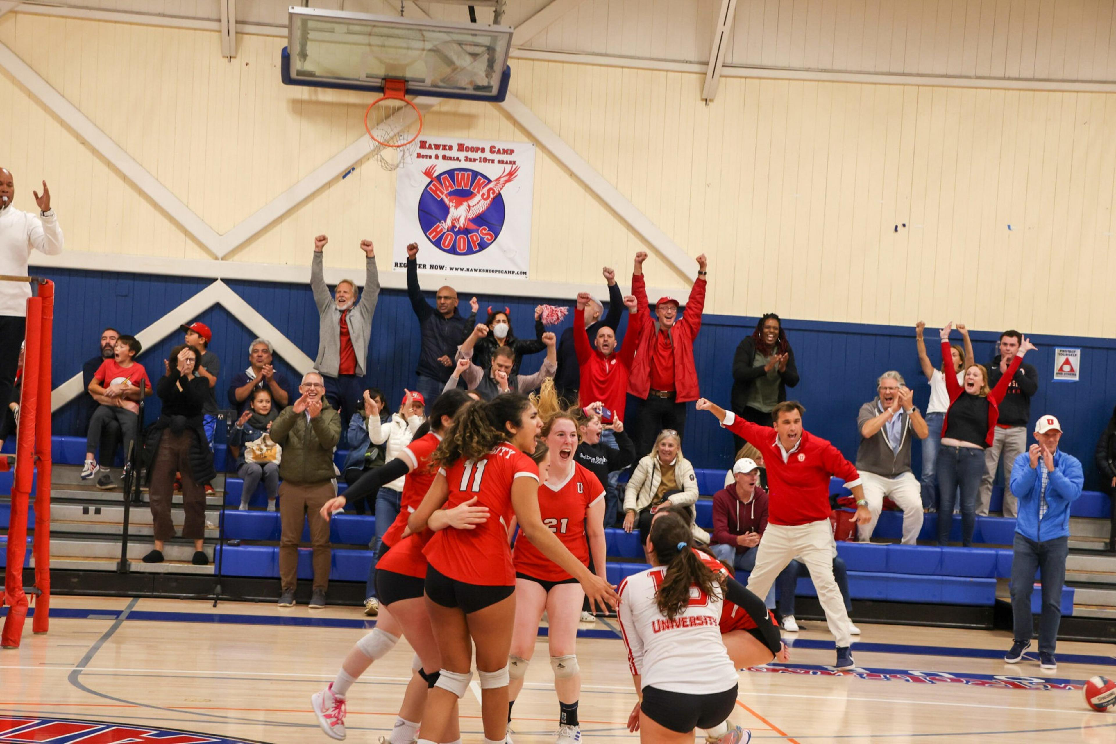 A volleyball team in red uniforms celebrates on the court while an excited crowd cheers loudly from the bleachers behind them.