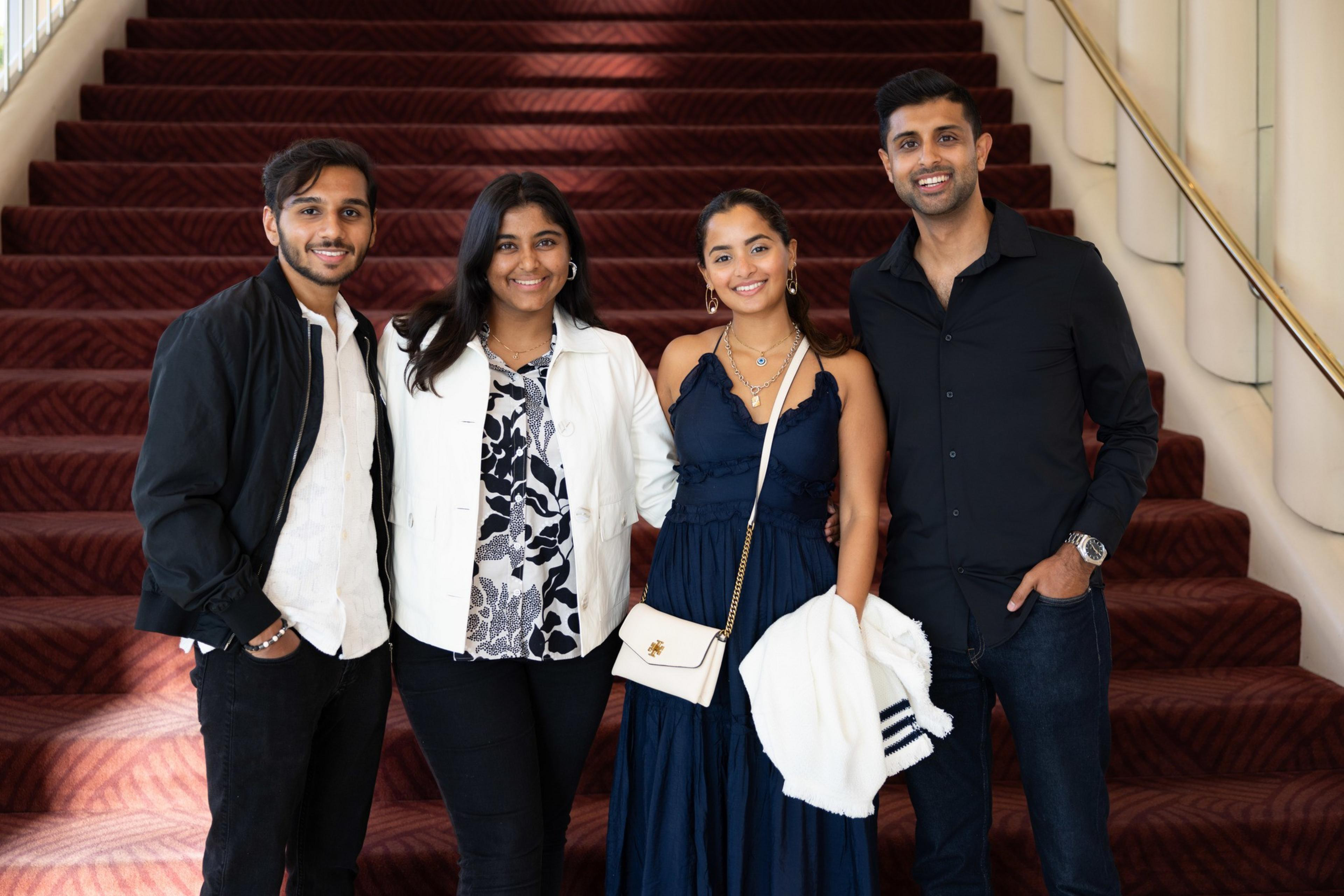 Four people stand on red-carpeted stairs, smiling. They are stylishly dressed: two men in black jackets and two women in elegant outfits, one holding a white purse.