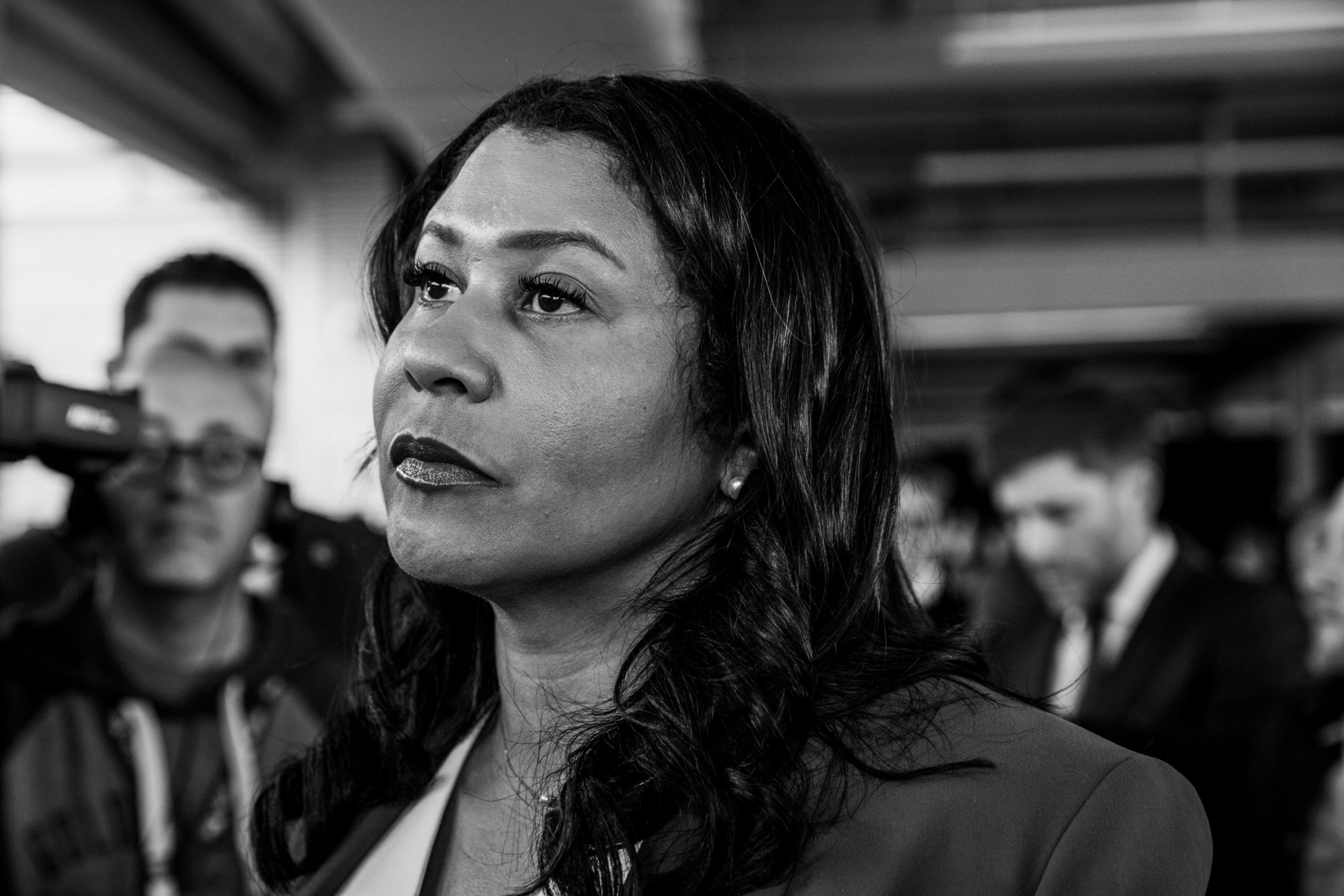 A black-and-white photo of a woman with a determined look, focused into the distance, with blurred figures in the background.