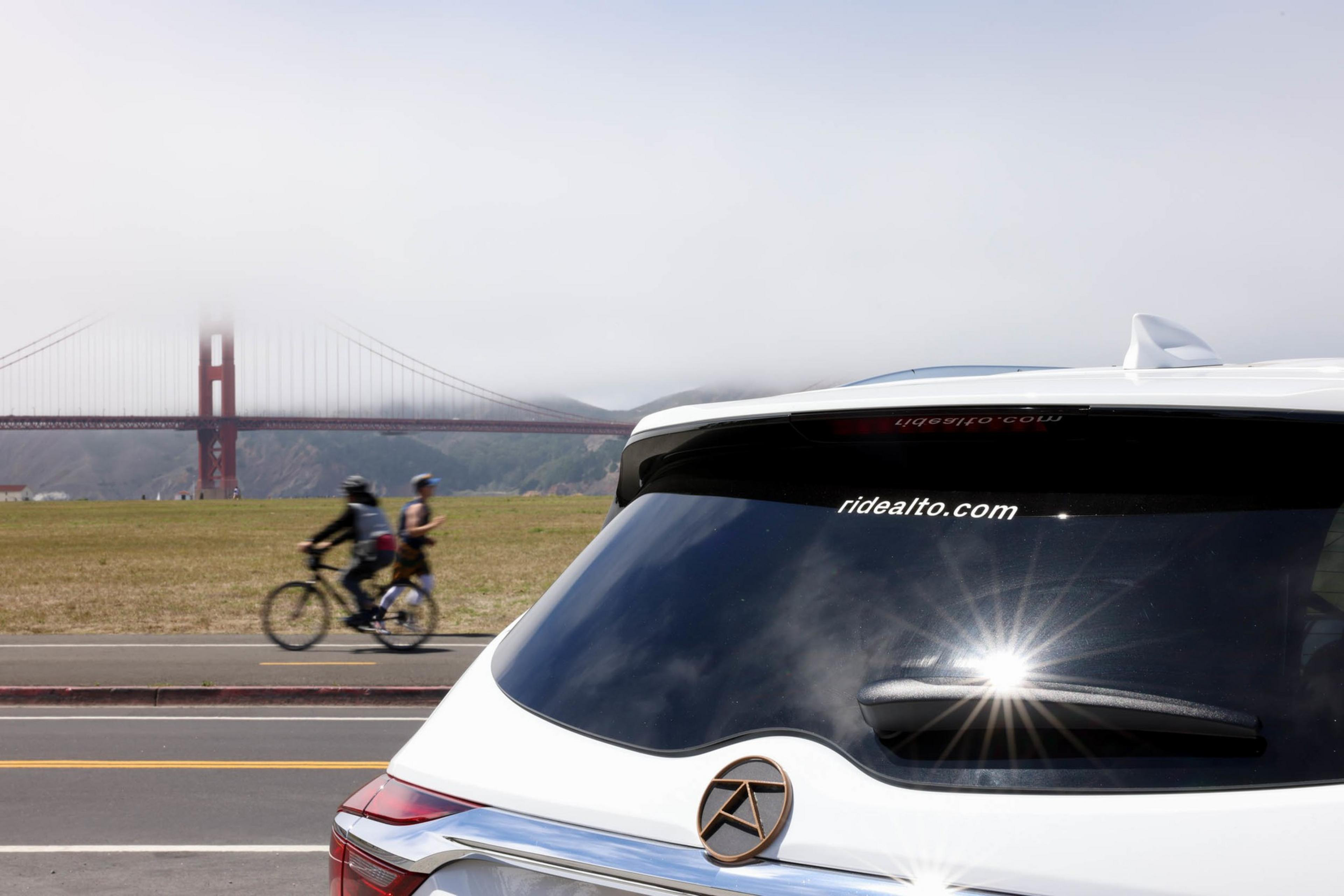 A white car with a logo and "ridealto.com" on its rear window is parked by a road where two cyclists ride, with the fog-covered Golden Gate Bridge in the background.